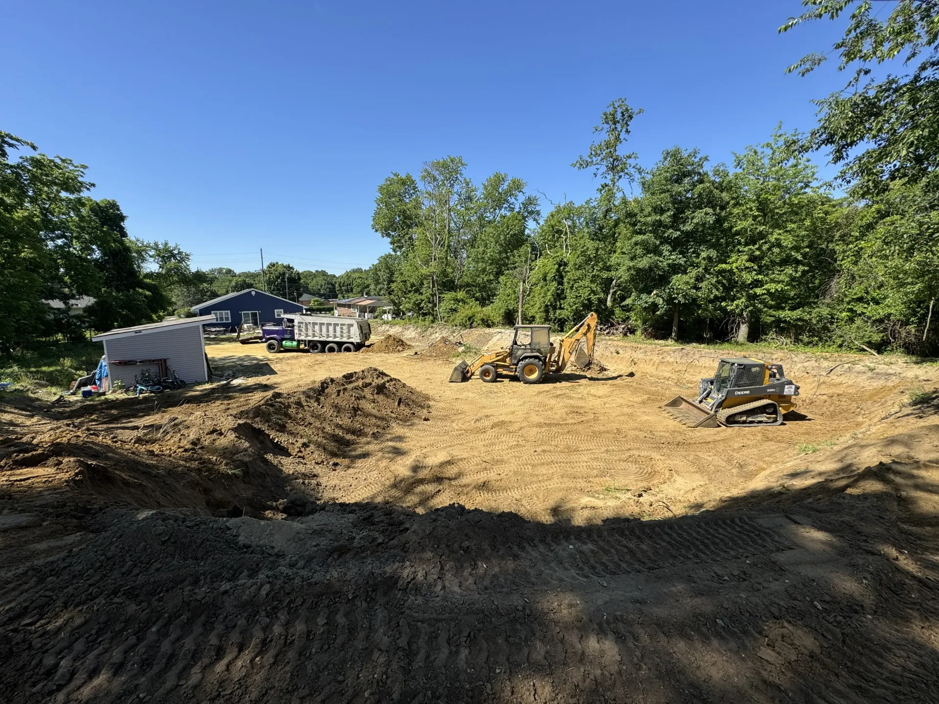 Dirt lot under construction with excavators, dump trucks, and piles of earth beneath a blue sky