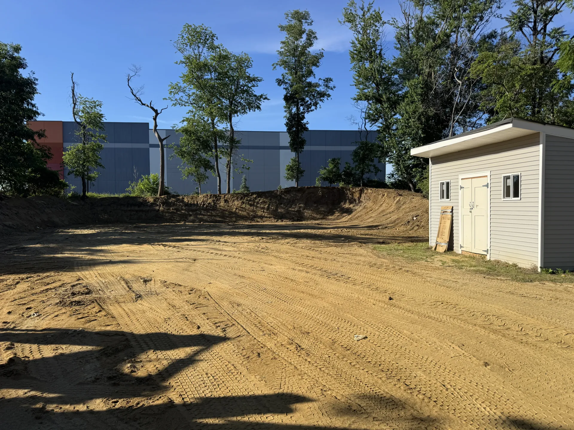 Dirt lot with trees and a small white shed beside a gray fence under a clear blue sky