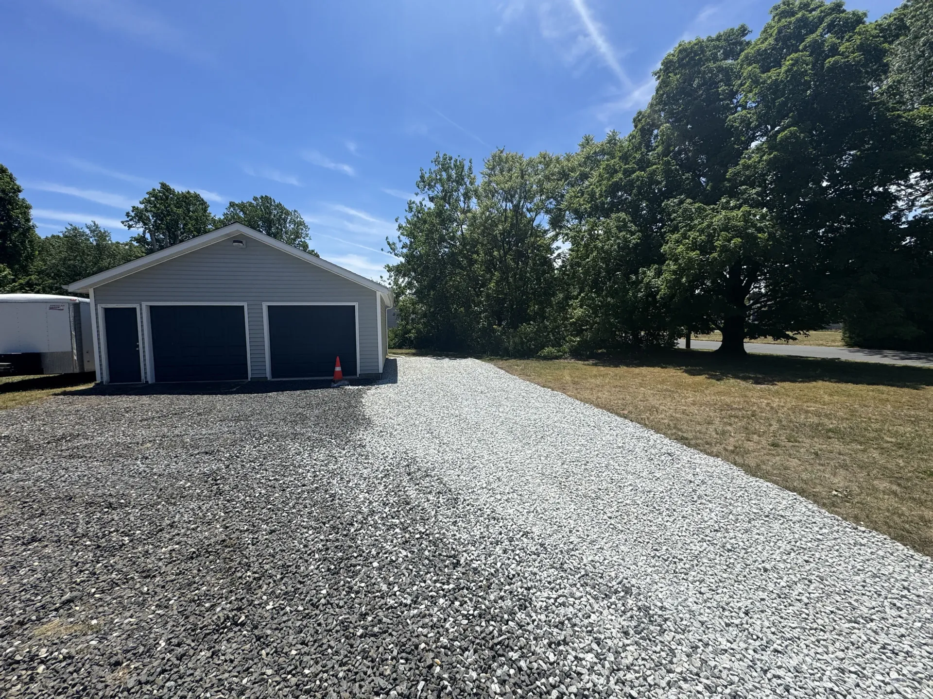 Gravel driveway leading to a white garage and trees under a bright blue sky