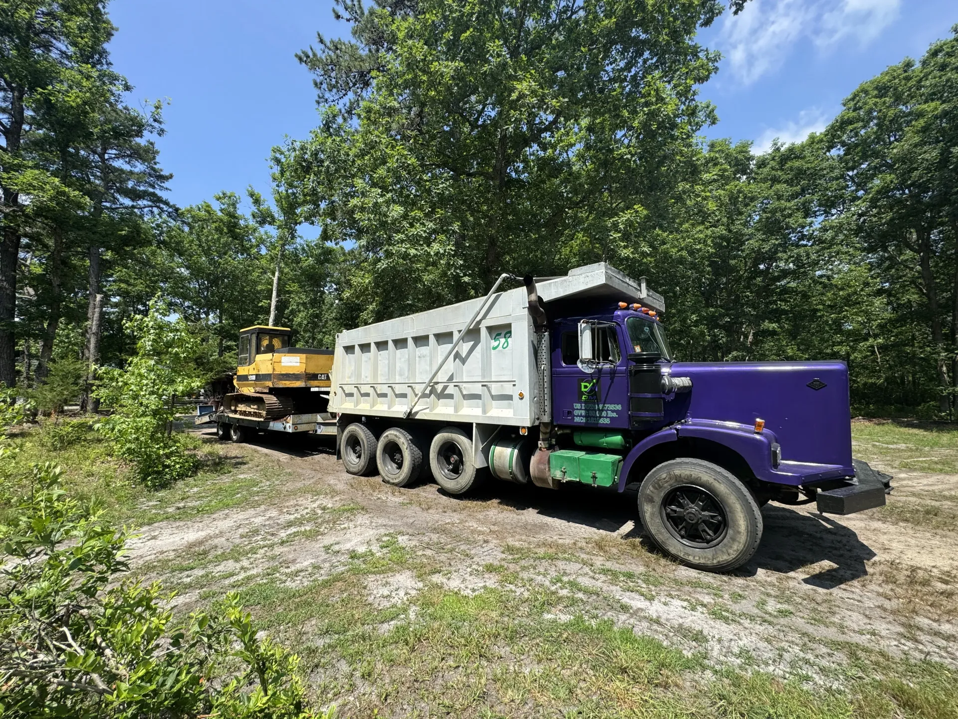 Purple dump truck with white trailer parked on a gravel road in a wooded area, with a yellow excavator behind it
