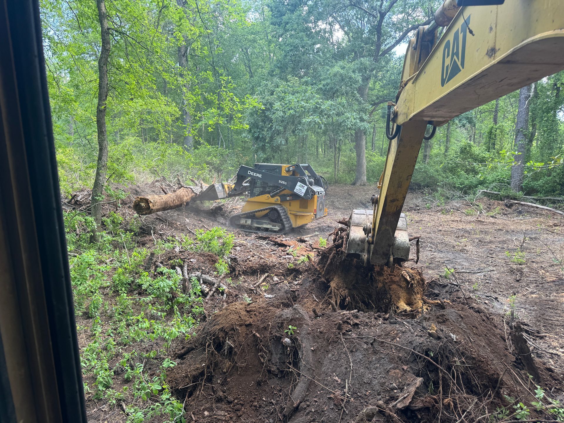 Excavator clearing a muddy wooded lot, with a skid steer in the background.