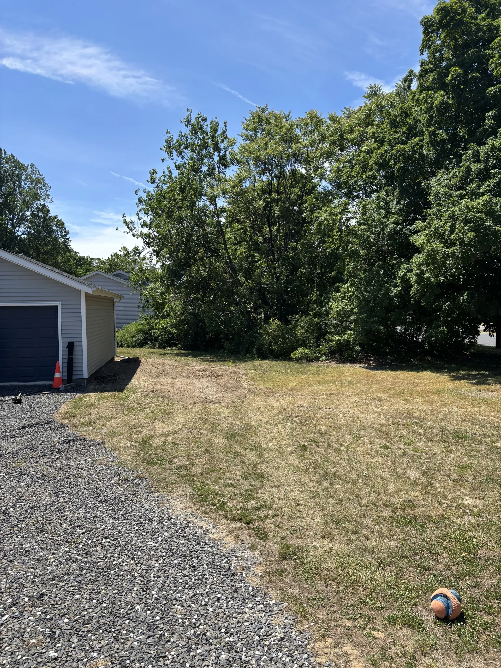 Gravel driveway beside a house with a grassy yard, large trees, blue sky, and a small ball in the foreground