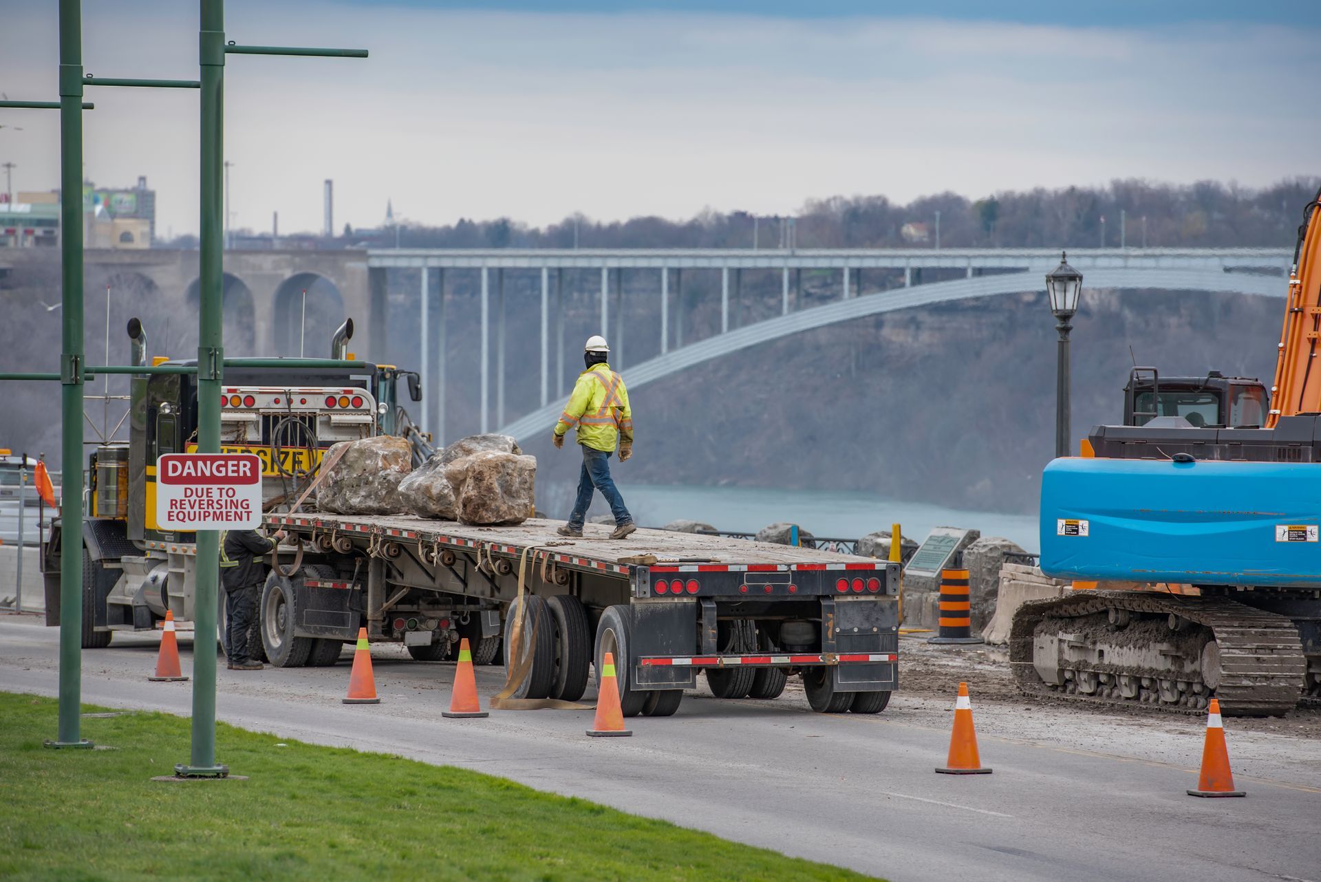 Construction worker on a flatbed truck carrying rocks near a bridge and excavator.
