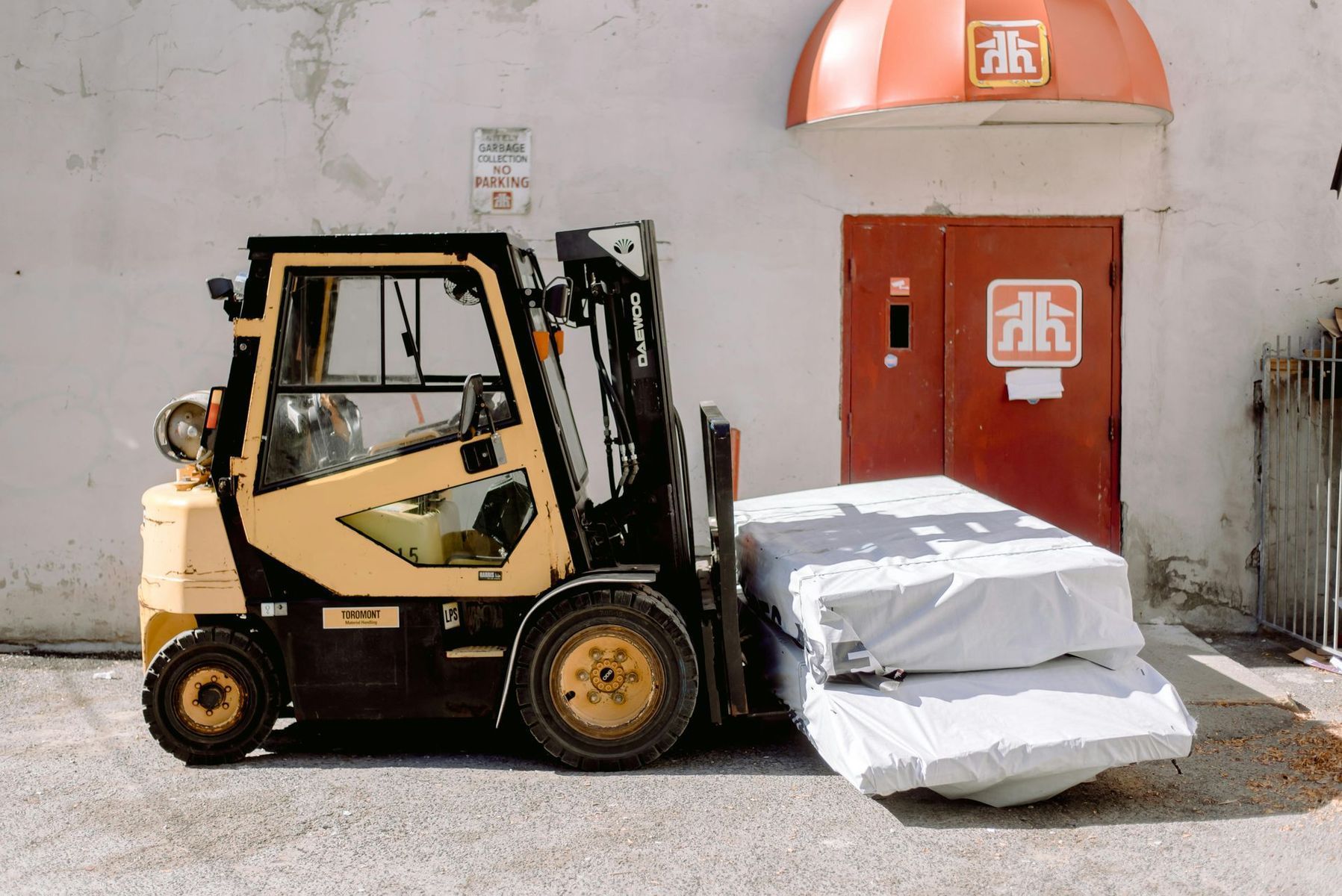 Yellow forklift carrying wrapped mattresses in front of a red-doored building.
