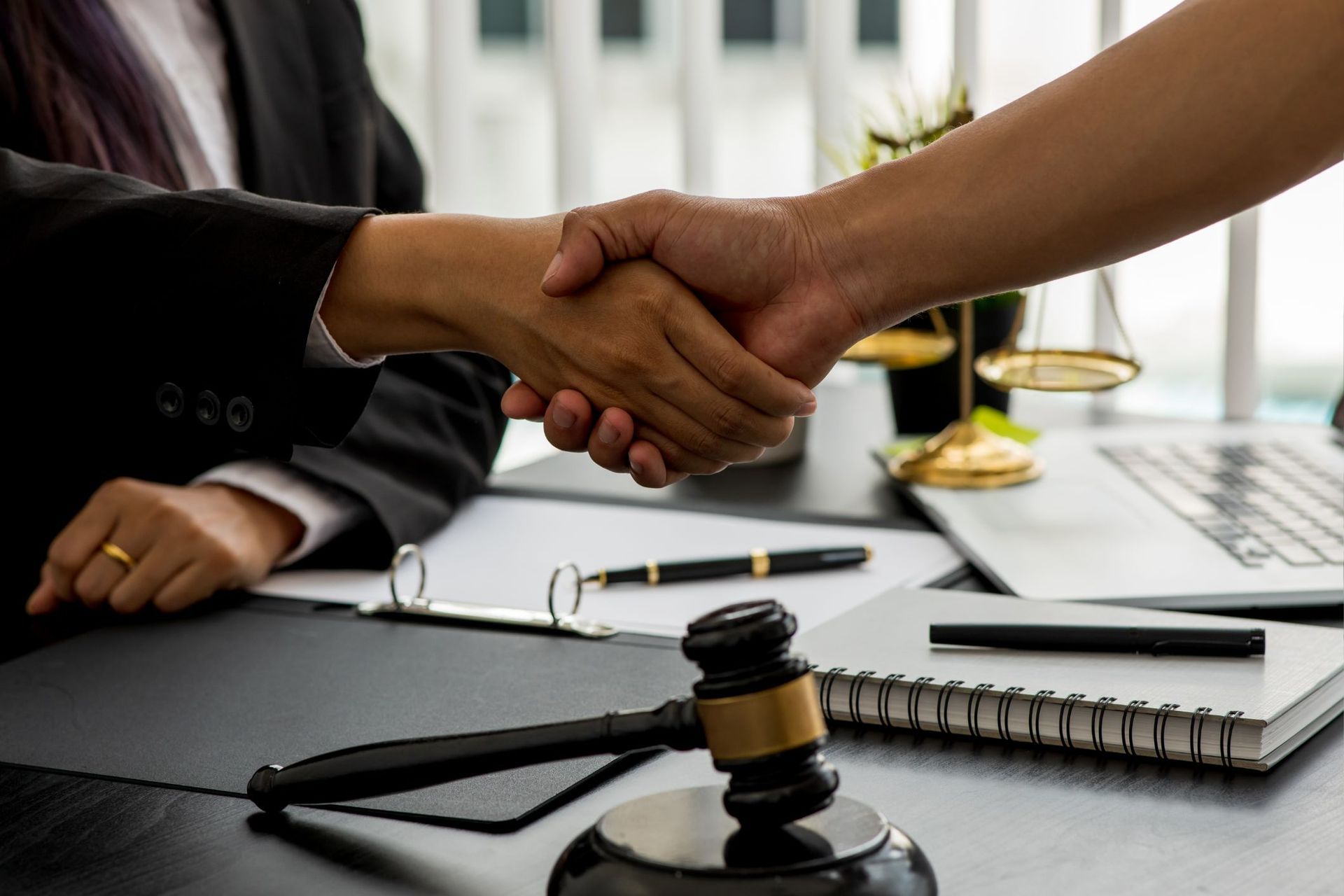 Handshake over a desk with legal documents, gavel, pen, and laptop in an office setting