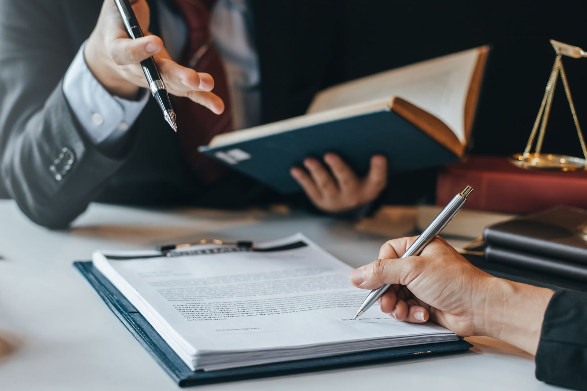Hands signing legal papers at a desk with a gavel, scales, and an open book nearby