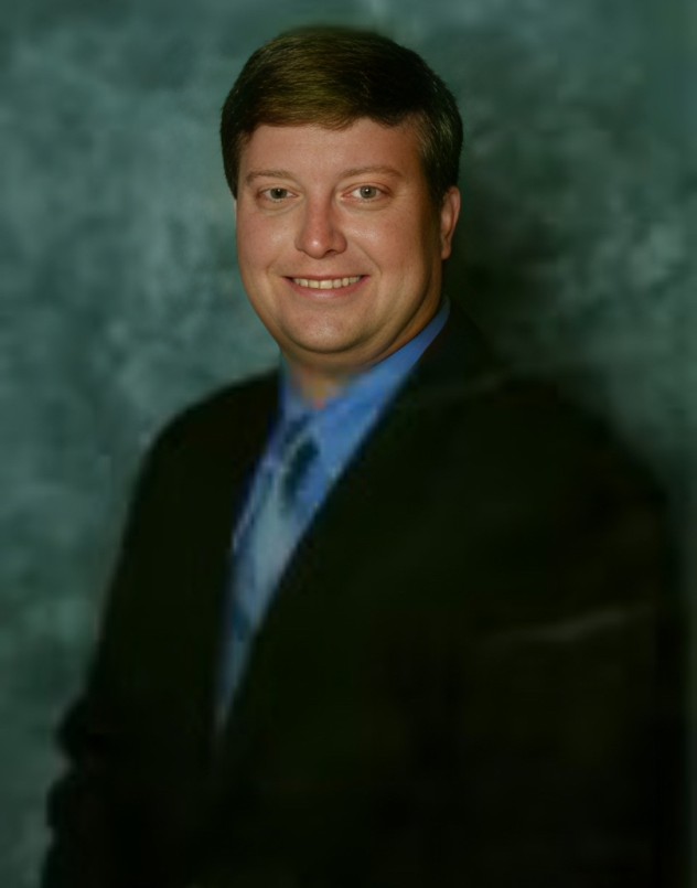 Smiling man in a black suit and blue shirt against a dark green studio backdrop