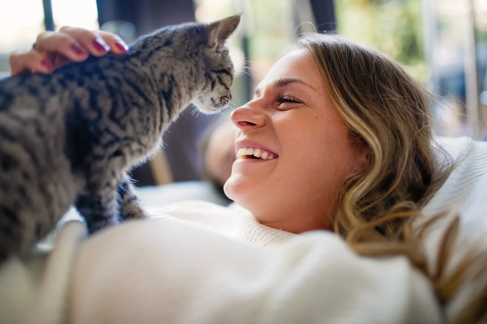 Woman lying on a couch smiles at a gray tabby cat, hand gently petting it.