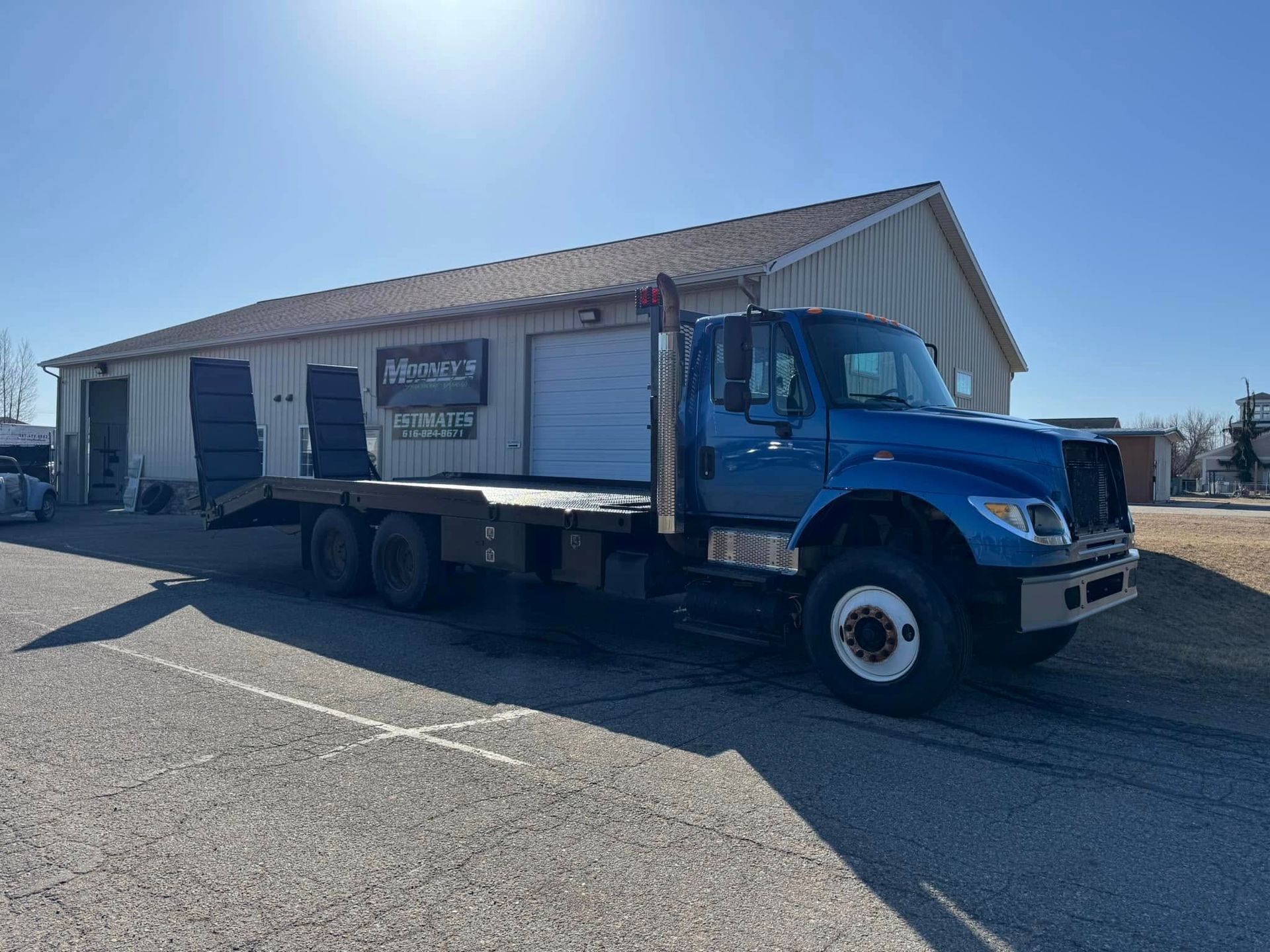 A blue flatbed tow truck parked on an asphalt lot in front of a metal warehouse building under a bright blue sky.