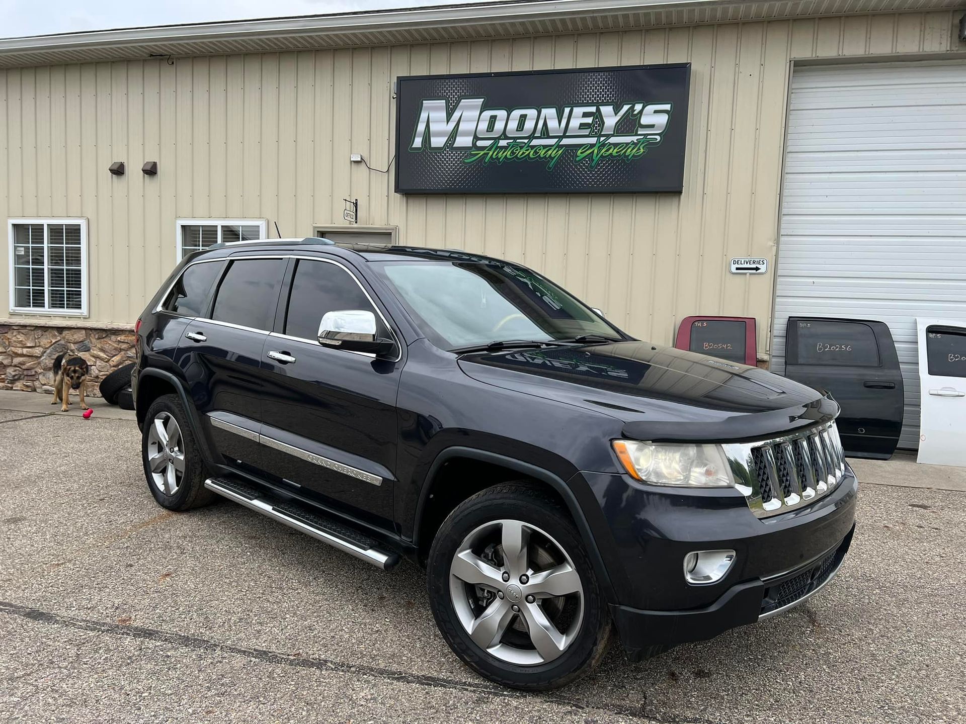 A black Jeep Grand Cherokee parked in front of a Mooney's automotive building on a gravel lot.