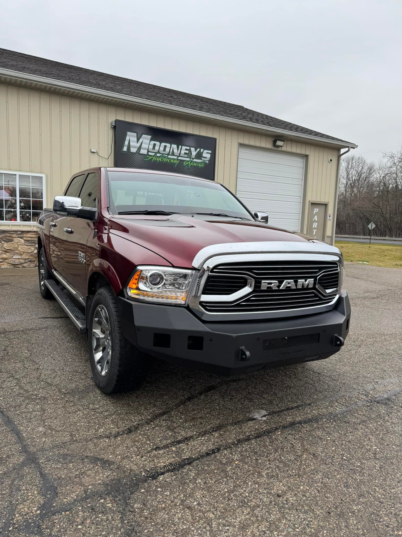 A maroon Ram pickup truck with a black aftermarket front bumper parked in front of a metal warehouse building.