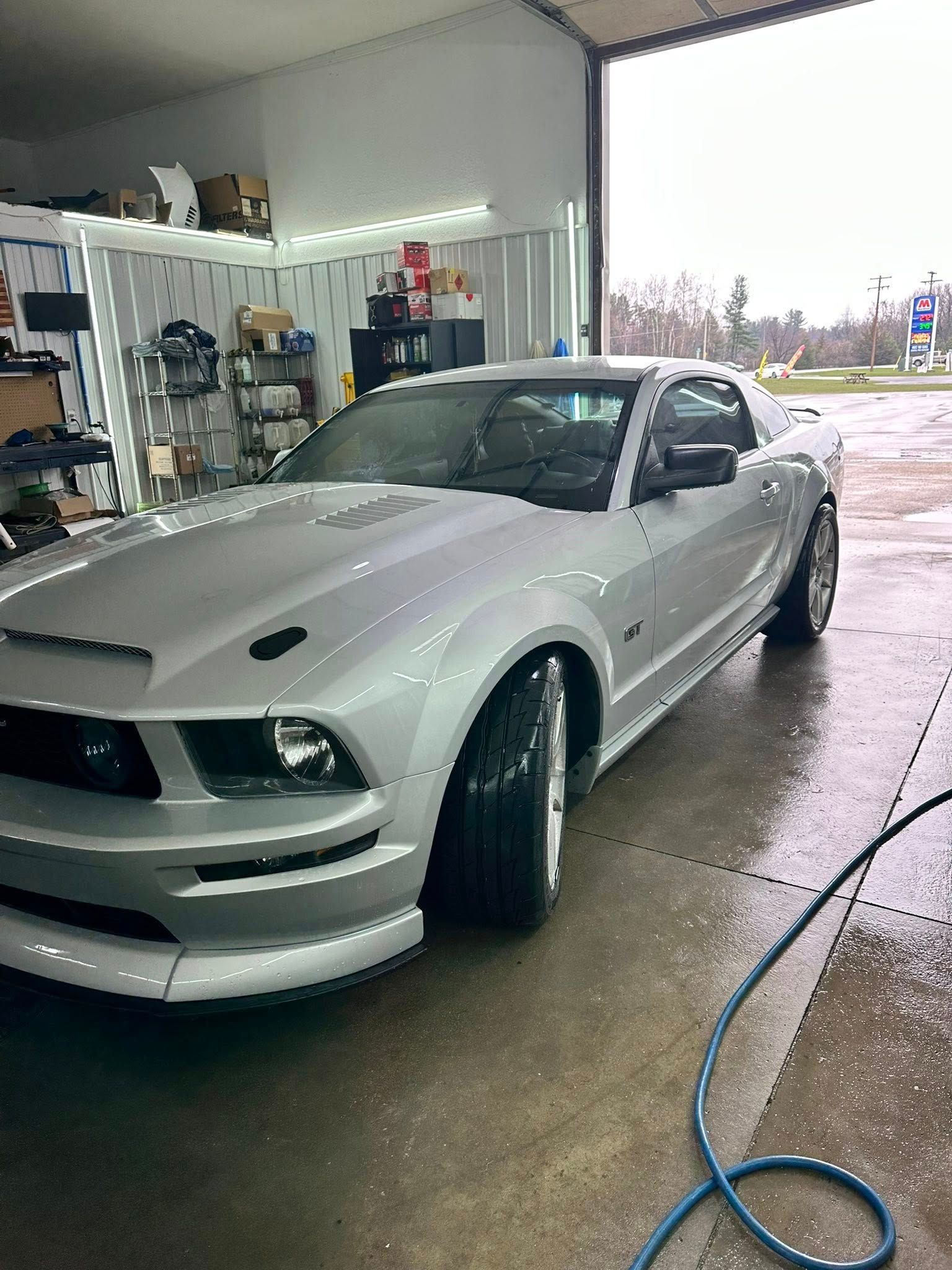 A silver Ford Mustang parked inside a garage with its hood pins visible.