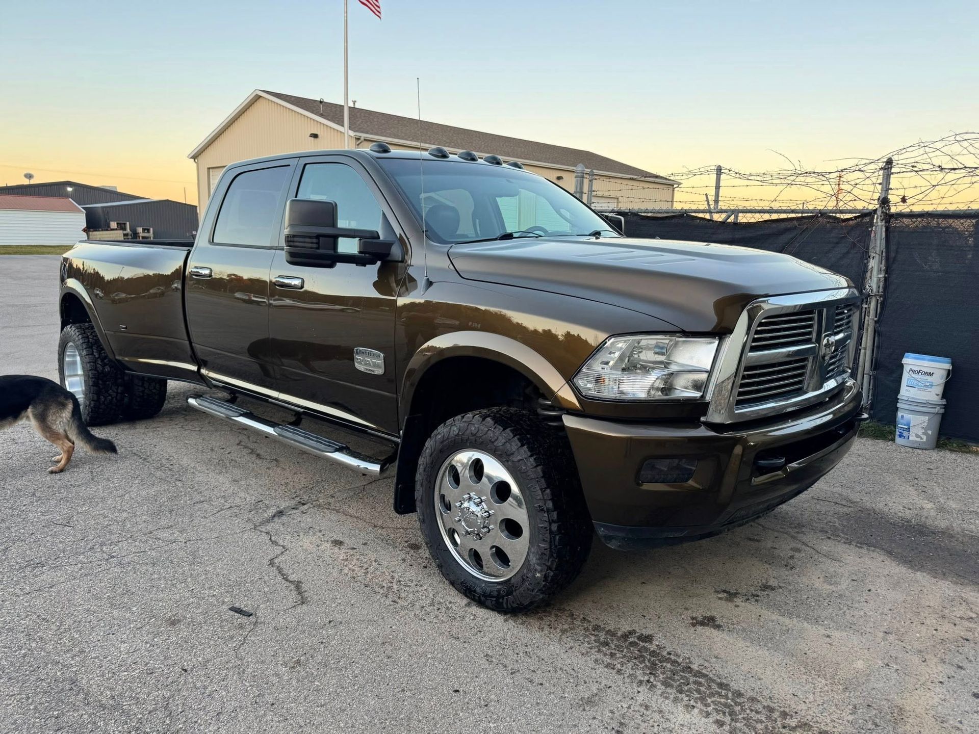 A dark brown Ram dually pickup truck parked in a gravel lot with a dog nearby.