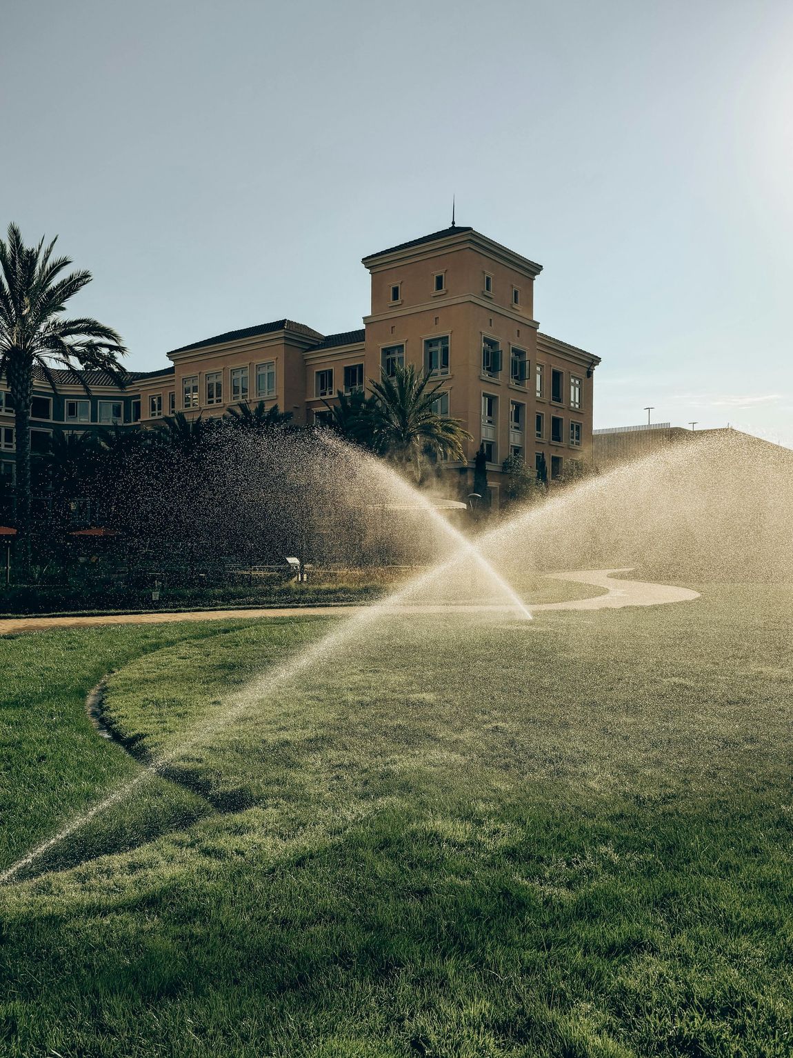 Lawn sprinkler spraying water in front of a tan building under a clear sky