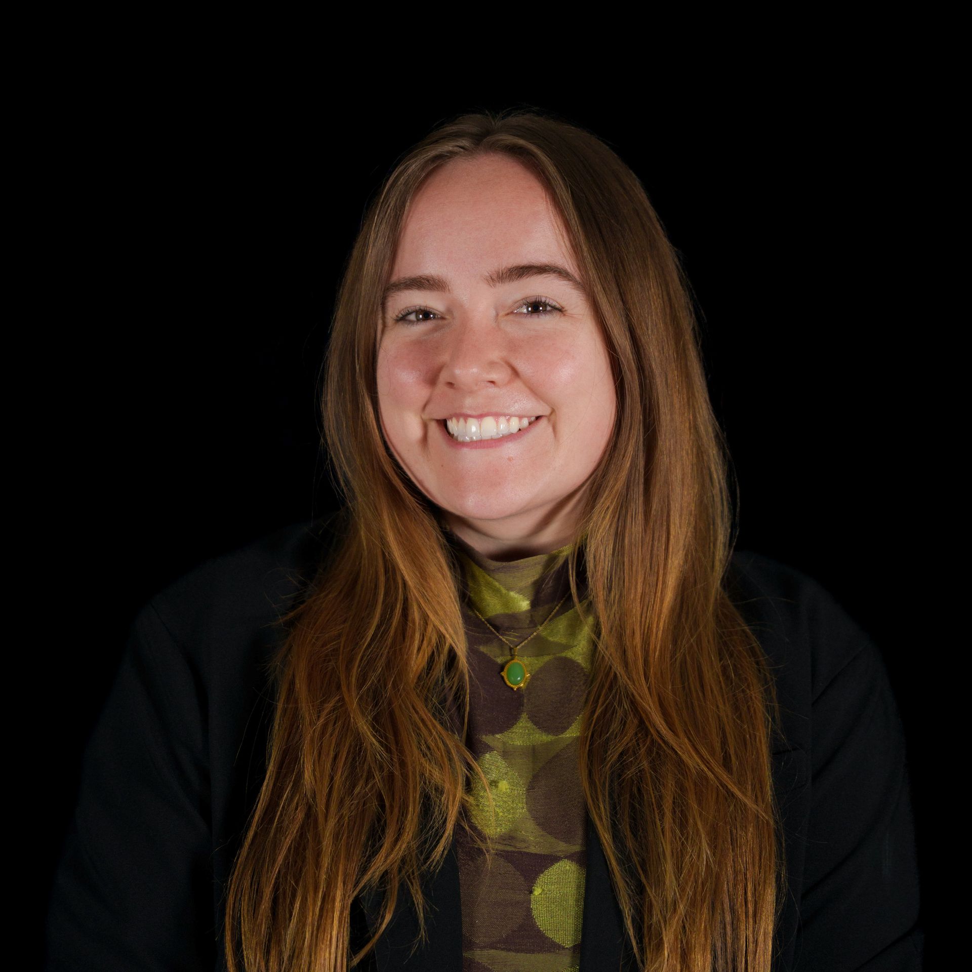 Woman with long brown hair smiles broadly against a black backdrop, wearing a dark blazer and hoop earrings.