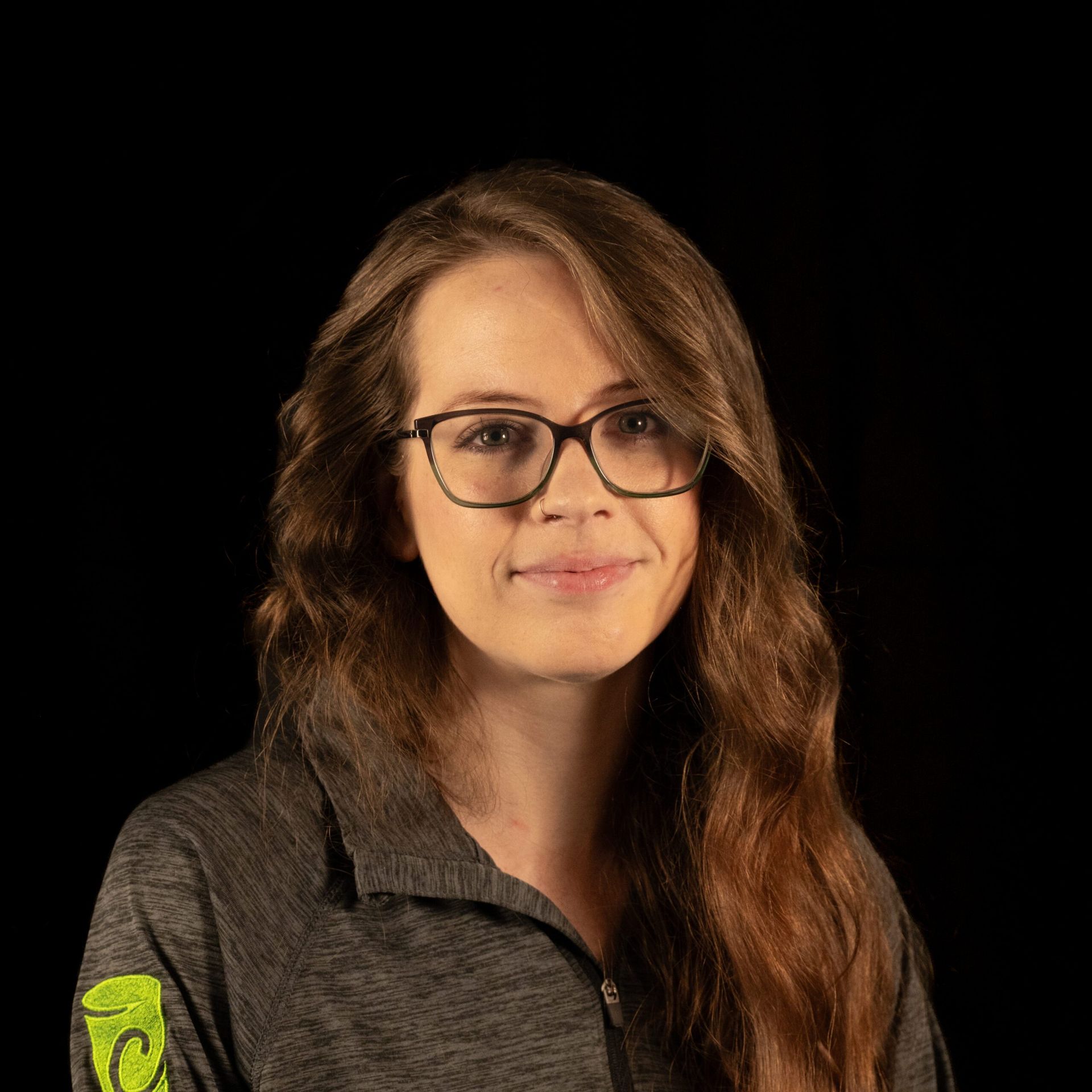 Woman with glasses smiling, wearing a gray zip-up shirt against a black background.