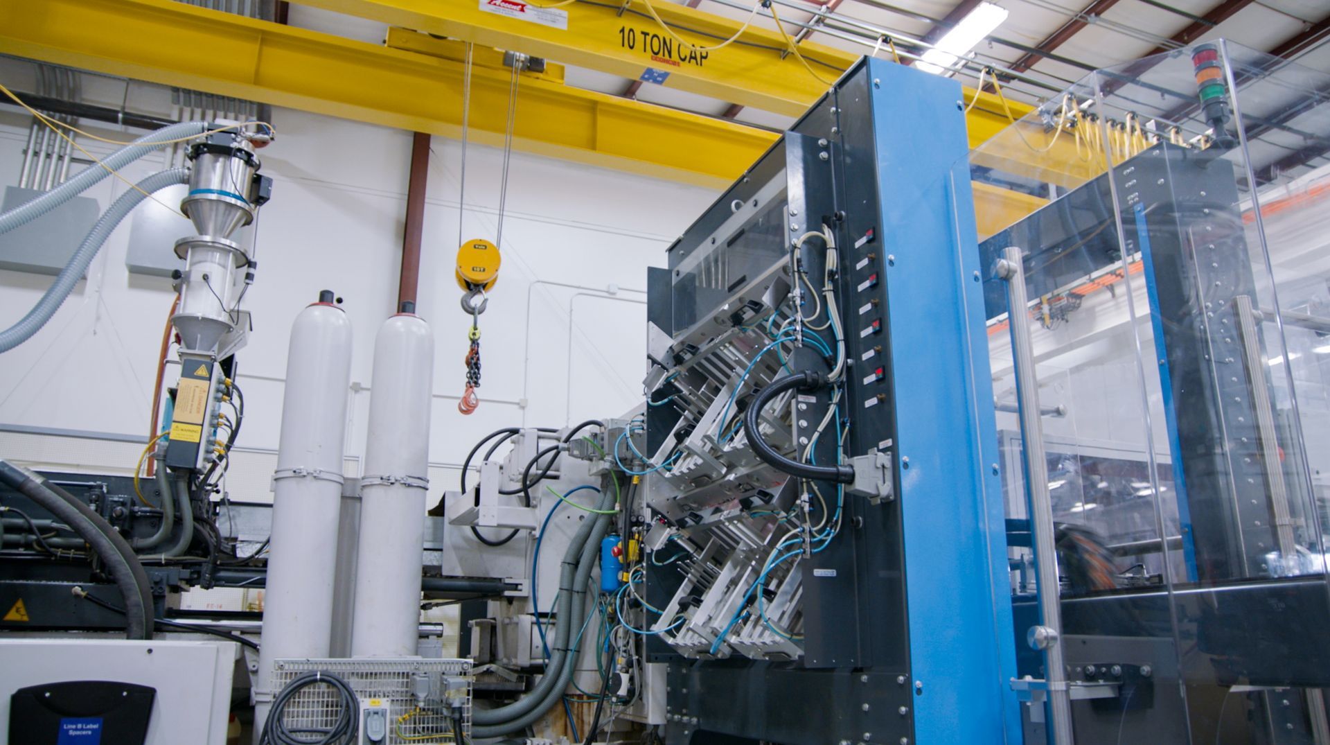Large blue and gray industrial machine in a factory, with yellow overhead beams and white pipes.