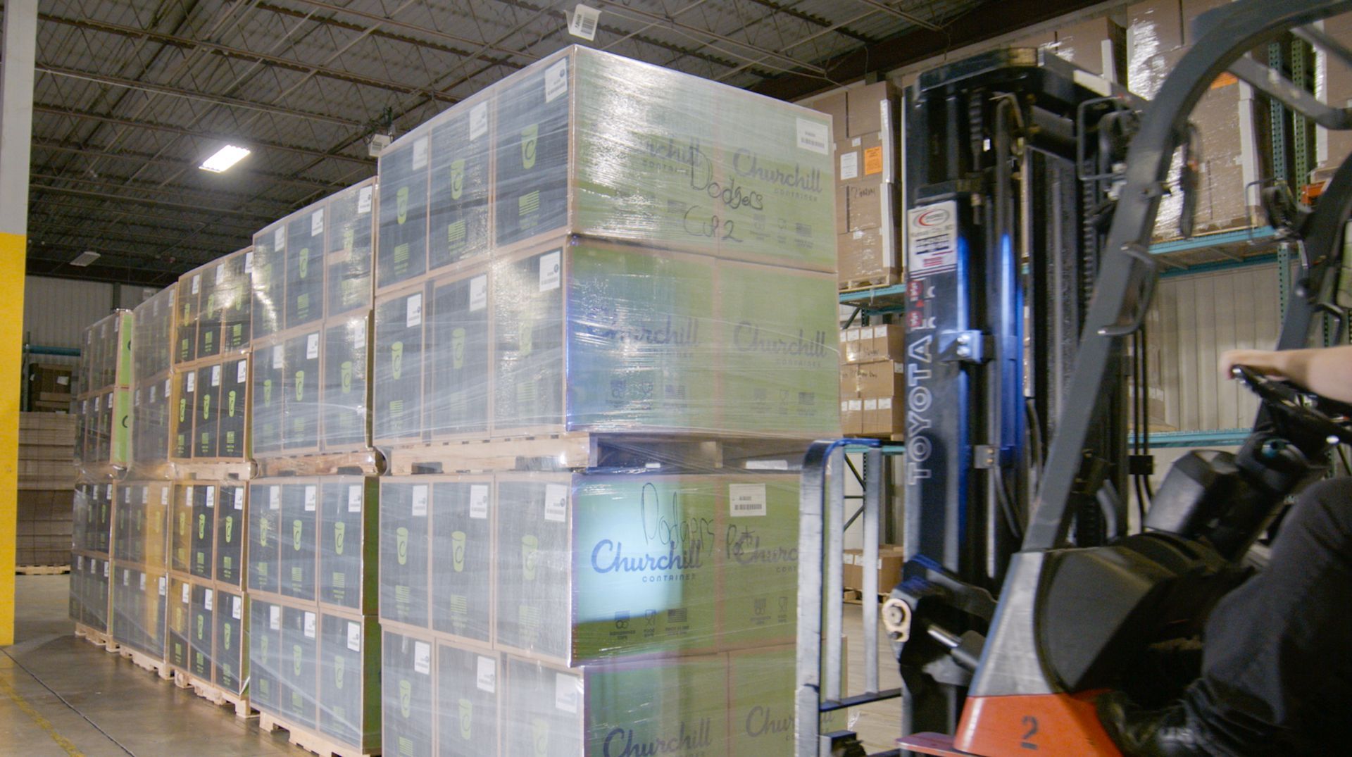 Forklift loading a stack of wrapped boxes in a warehouse, boxes are arranged in rows.