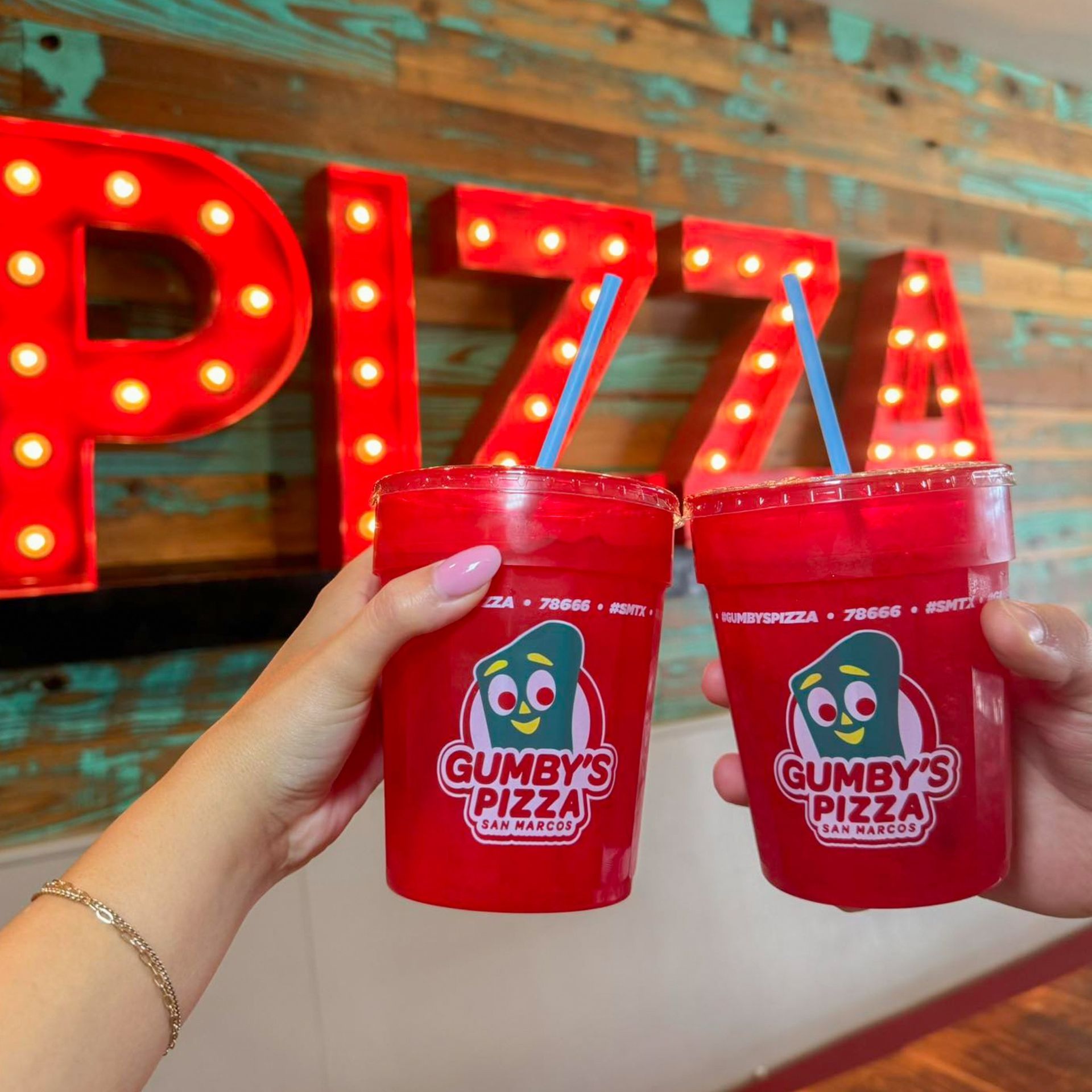 Hands holding two red Gumby's Pizza drinks in front of a pizza sign.