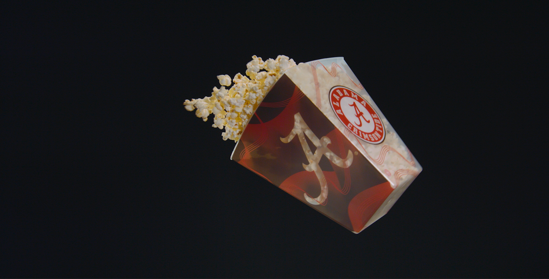 Popcorn box with Alabama logo spilling popcorn, black background.