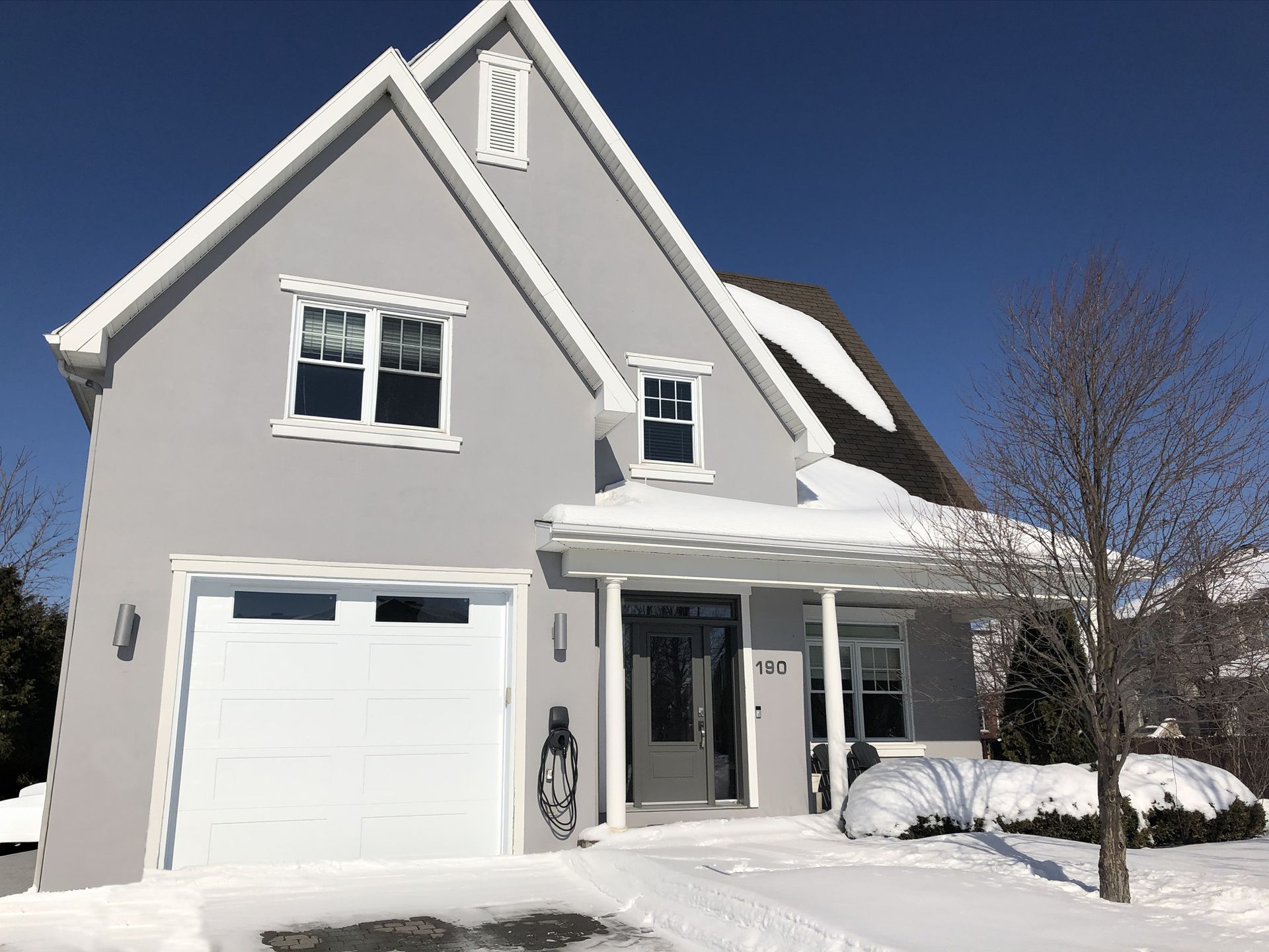 Snow-covered gray house with attached garage, white trim, and a front porch under a clear blue sky