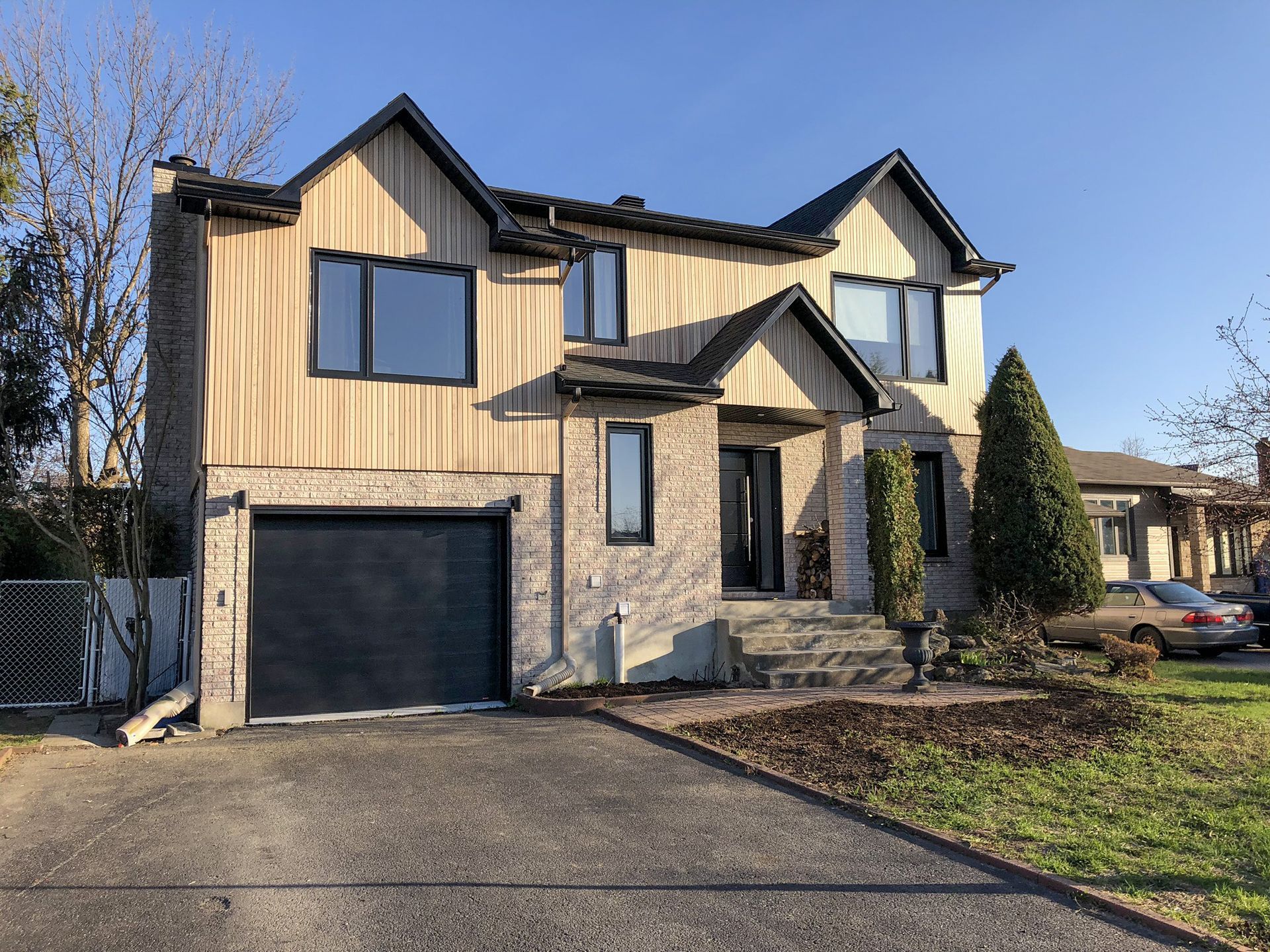 Two-story tan house with black garage door and stone accents beside a driveway and lawn.