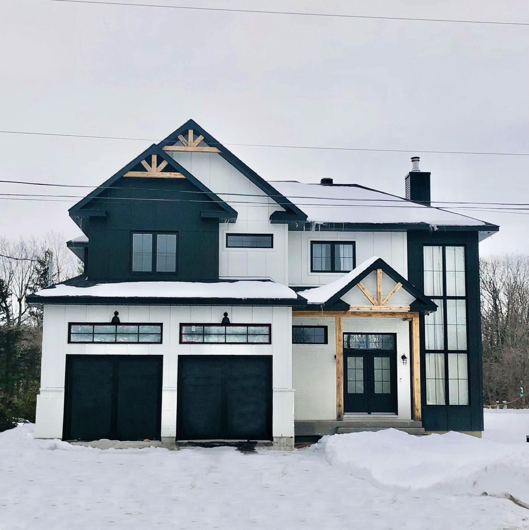 Snow-covered modern two-story house with dark trim and a double garage under an overcast sky