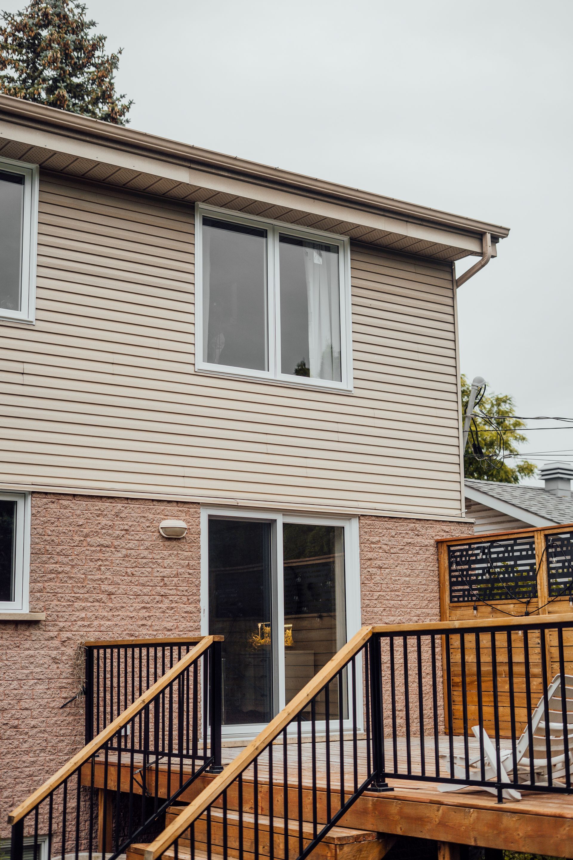 Maison beige à deux étages avec terrasse en bois, rambarde noire et porte coulissante en verre