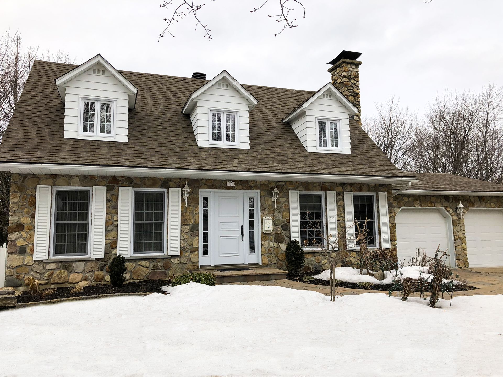 Maison en pierre enneigée, avec des volets blancs et trois lucarnes, sous un ciel nuageux