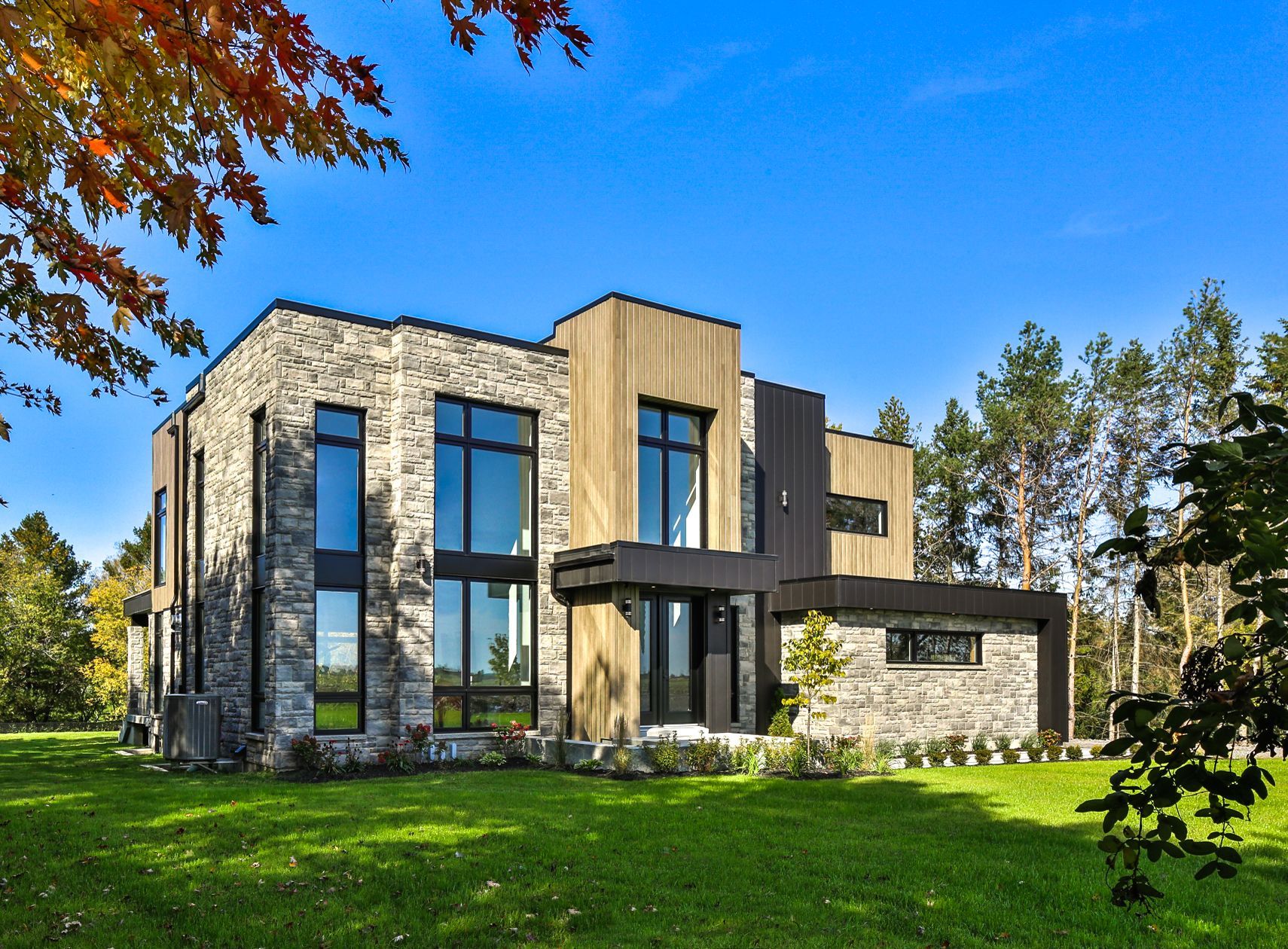 Maison moderne en pierre et en bois avec de grandes fenêtres, située sur une pelouse verdoyante sous un ciel d'un bleu éclatant.