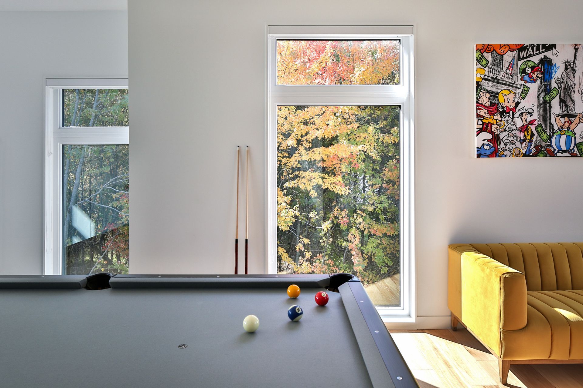 Salon lumineux avec table de billard, grandes fenêtres, arbres d'automne, canapé jaune et décorations murales colorées.