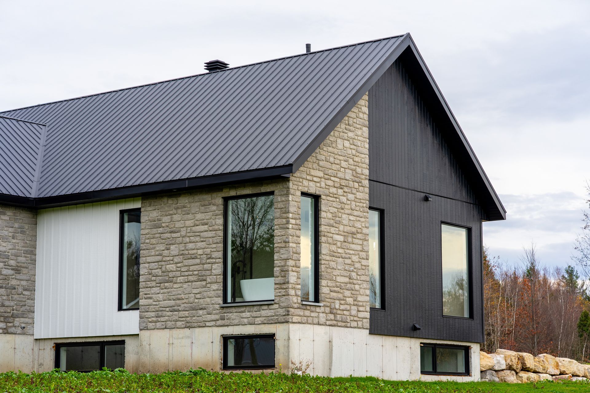 Maison moderne avec toit en métal foncé, revêtement en pierre et noir, et grandes fenêtres dans un cadre rural