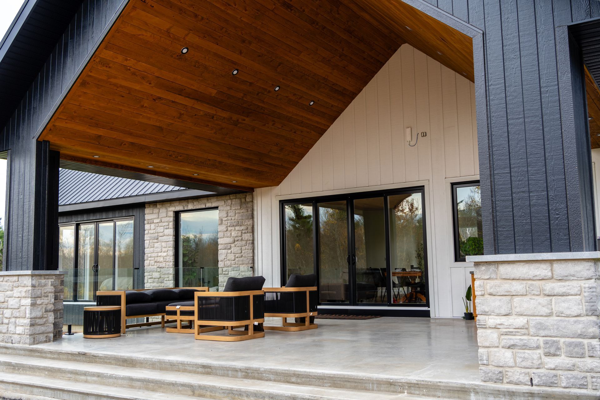 Covered patio with wood ceiling, stone columns, black chairs, and large glass doors.