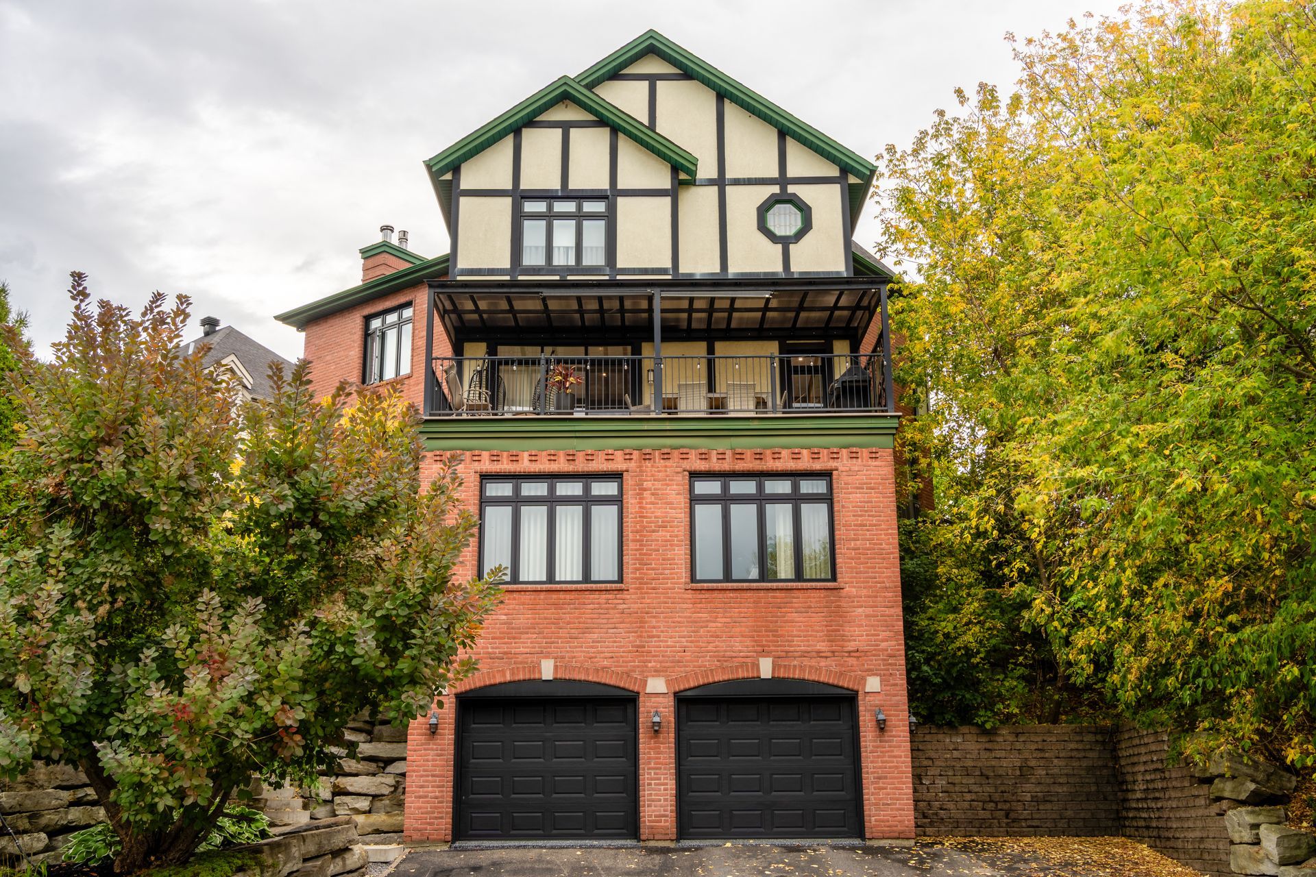 Brick house with green trim, a balcony, and two black garage doors surrounded by trees.