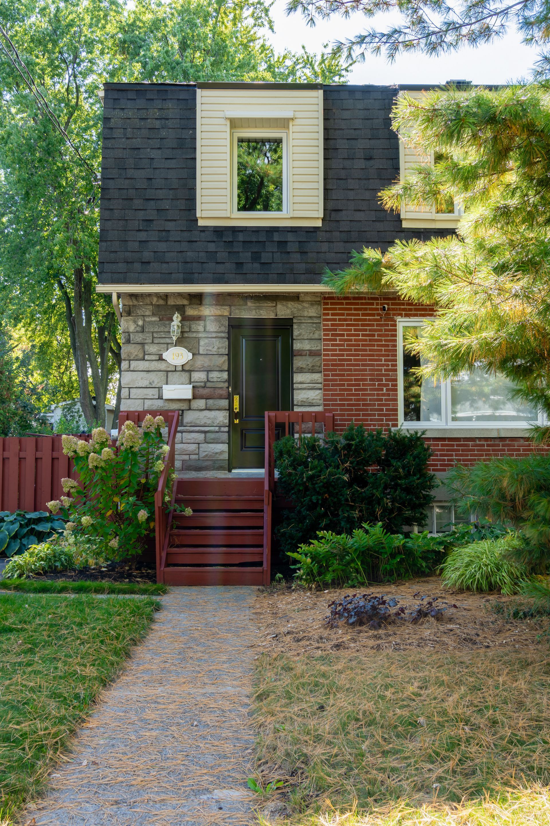 Petite maison de deux étages en pierre et en bardeau, avec marches rouges, allée d'entrée et jardin arboré.