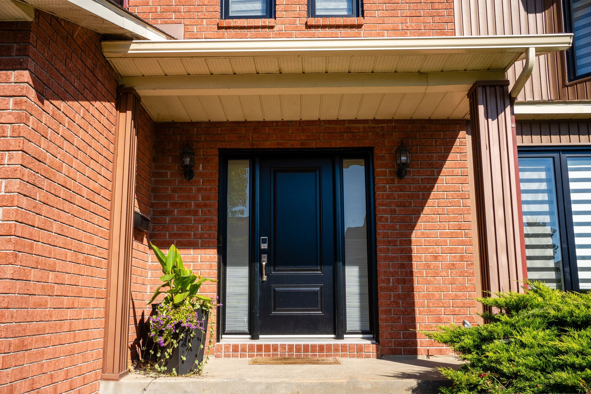 Front entrance of a brick townhouse with a black door, small potted plant, and concrete steps.