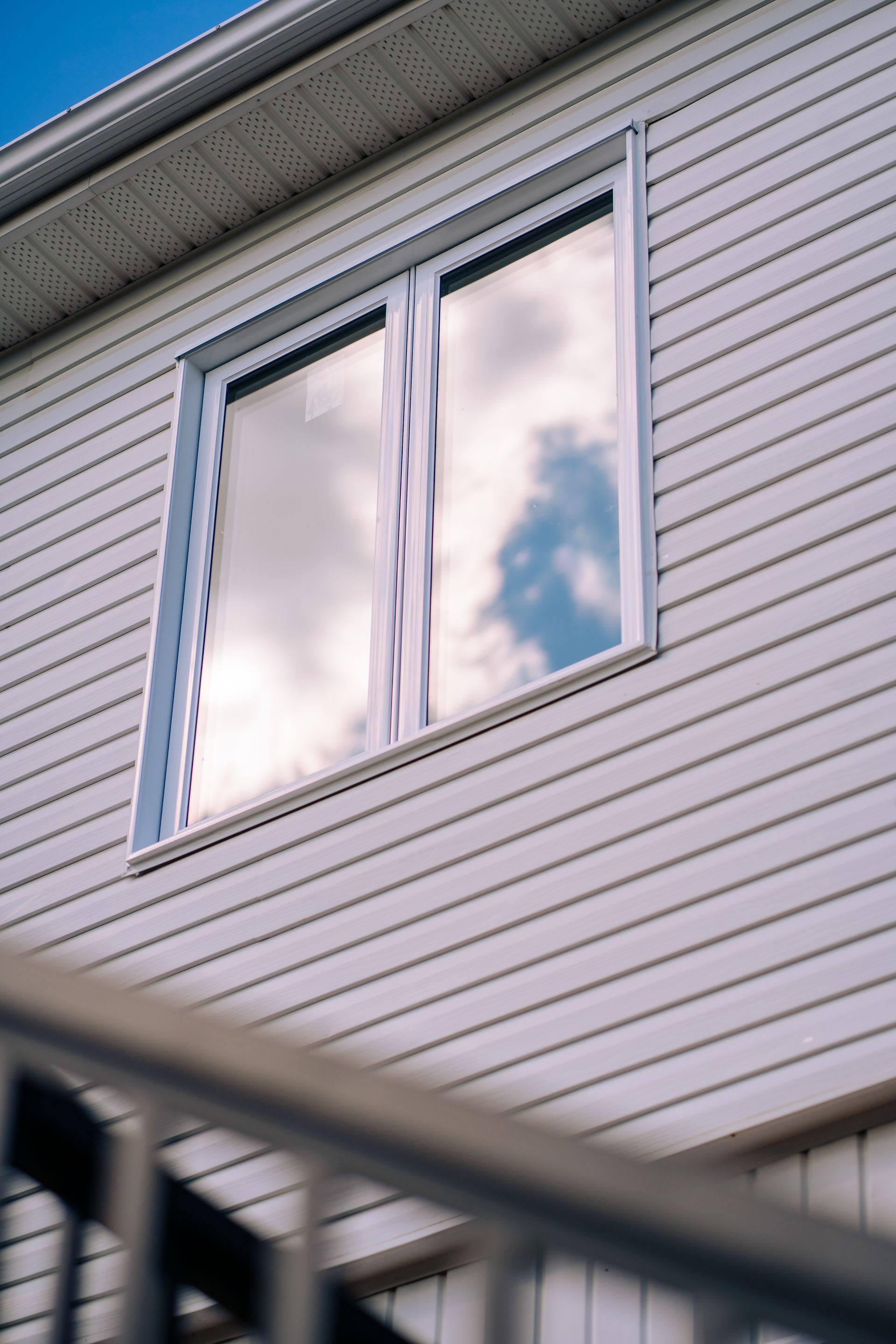 Façade blanche d'une maison avec une fenêtre au deuxième étage reflétant les nuages, vue par-delà une balustrade sombre.