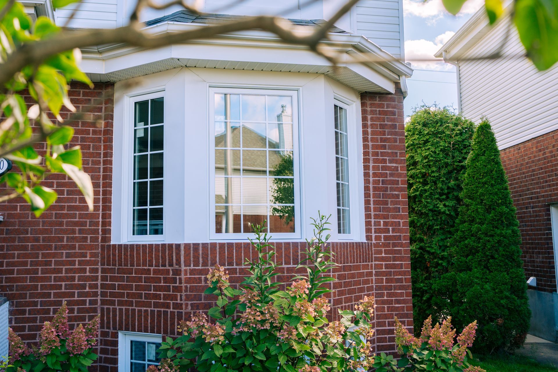 Brick house exterior with white bay window, red shutters, and green shrubs in front
