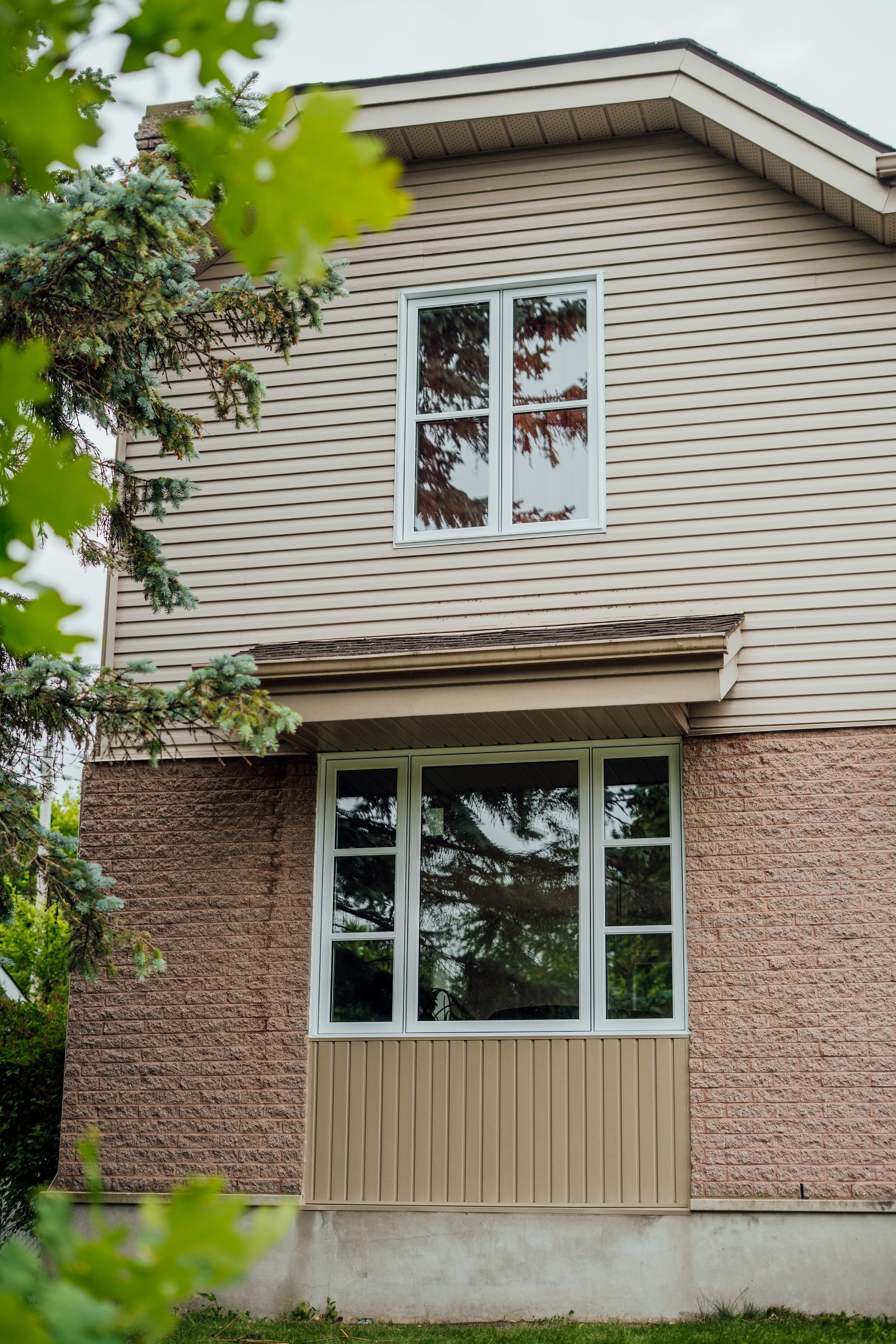 Two-story house exterior with beige siding, brick lower wall, and white-framed windows