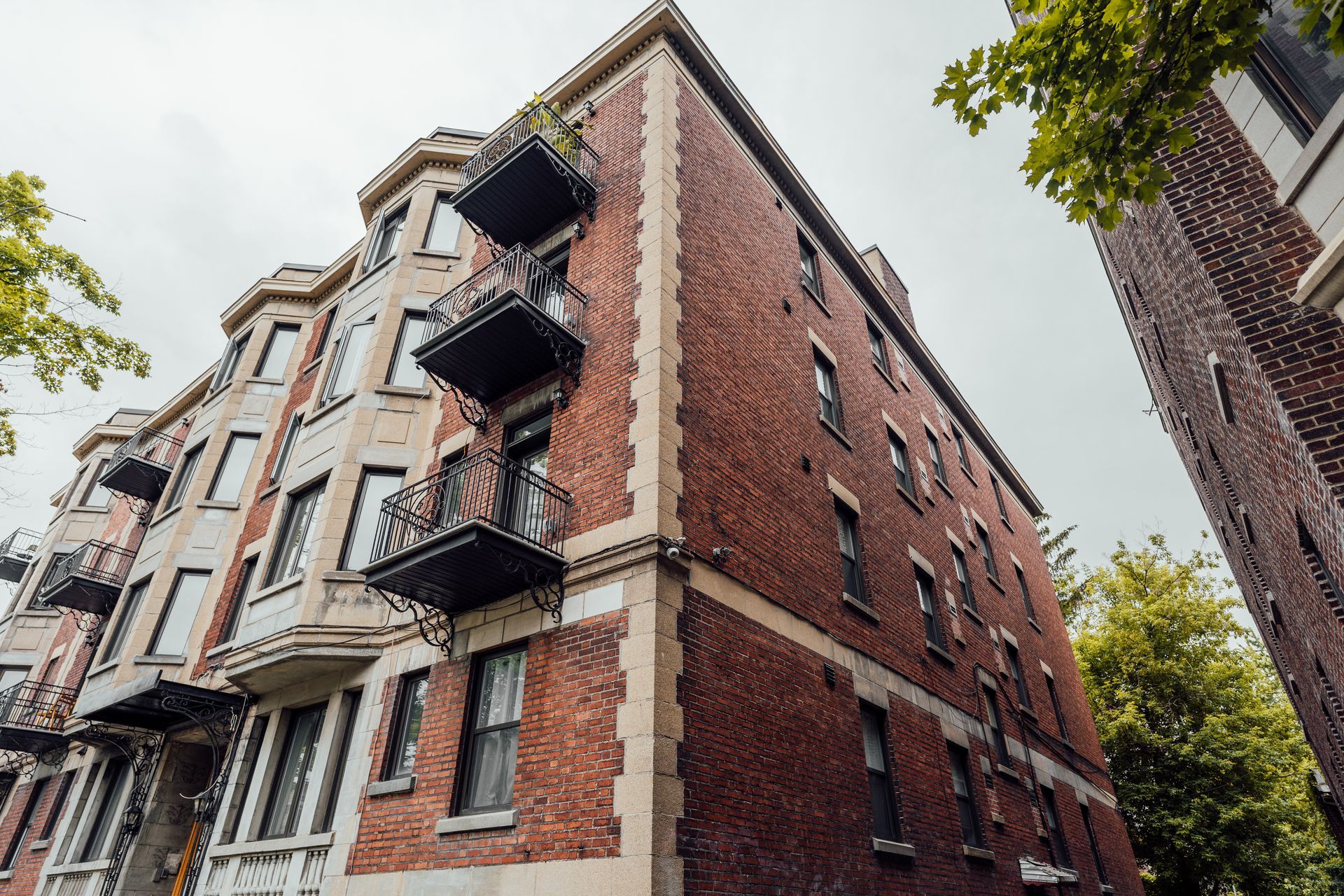 Red brick apartment building with black fire escapes viewed from below on a tree-lined street