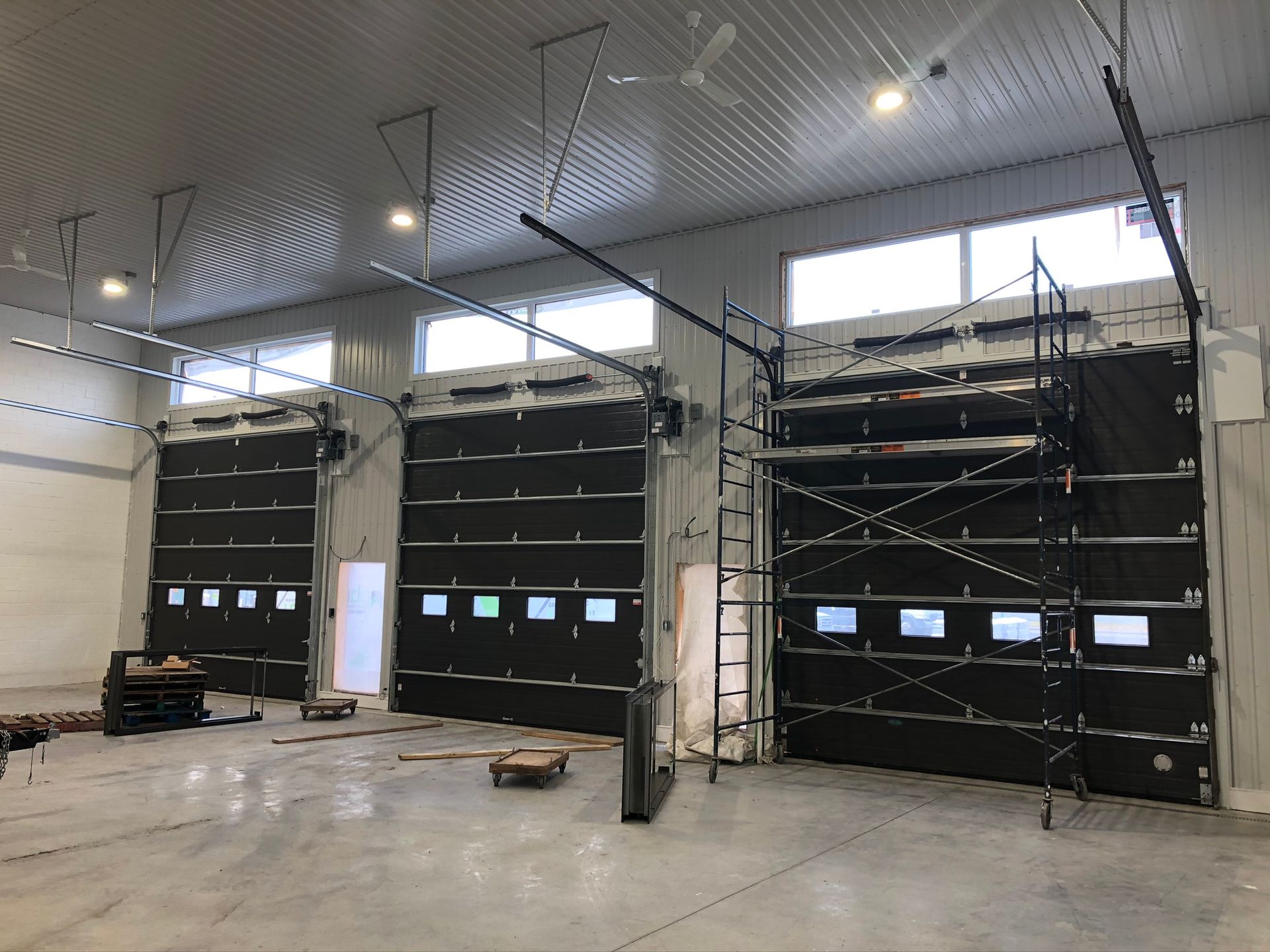 Grand atelier industriel avec trois portes sectionnelles noires, échafaudages et éclairage zénithal puissant.