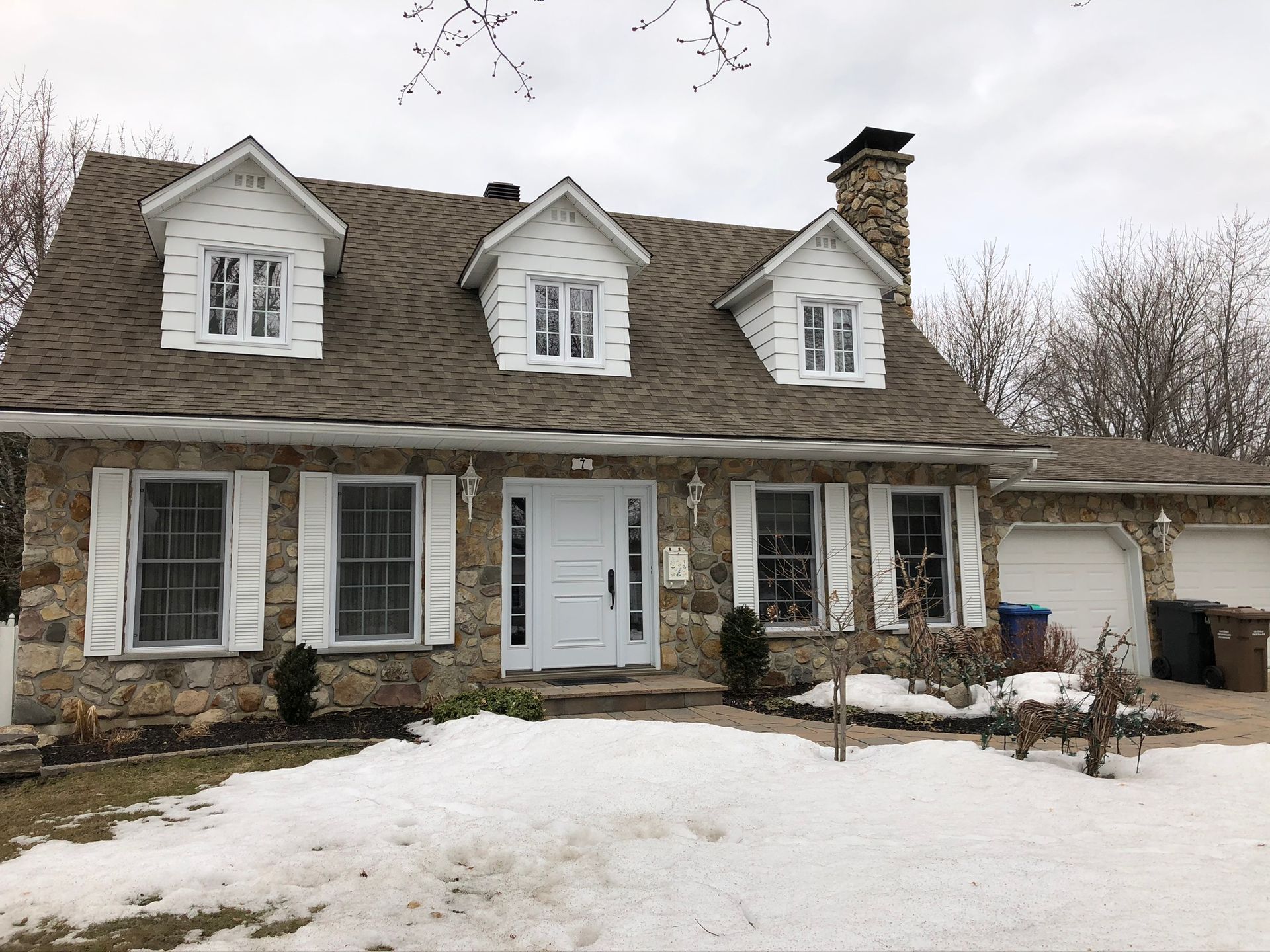 Snow-covered suburban house with dormer windows, stone facade, and a garage under a cloudy sky