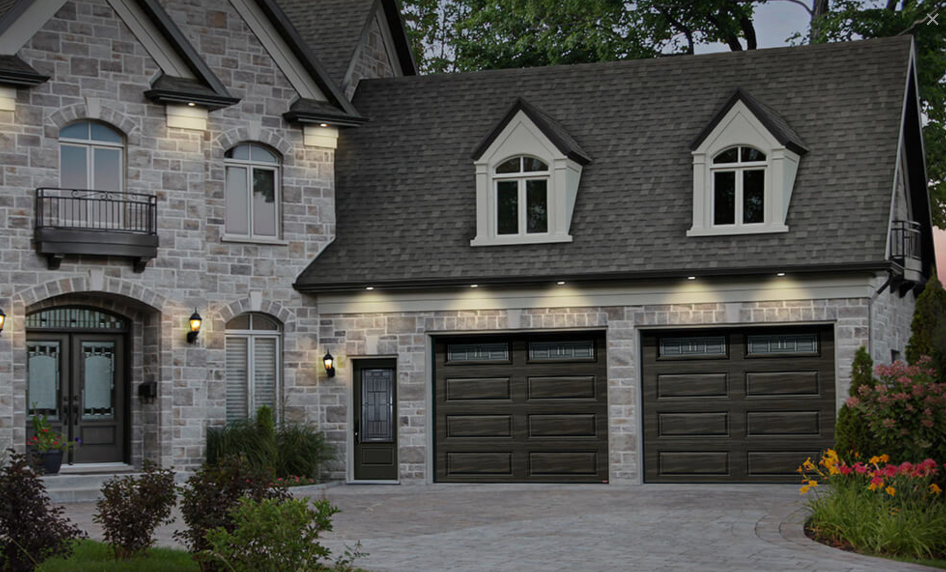 Stone house with dark roof, two garage doors, dormer windows, and lit exterior entry at dusk