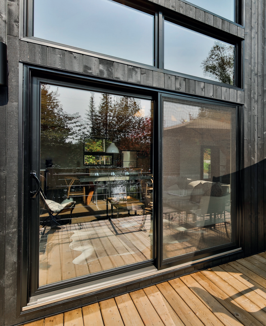 Porte vitrée moderne à cadre noir reflétant une terrasse ensoleillée avec terrasse en bois et mobilier de jardin