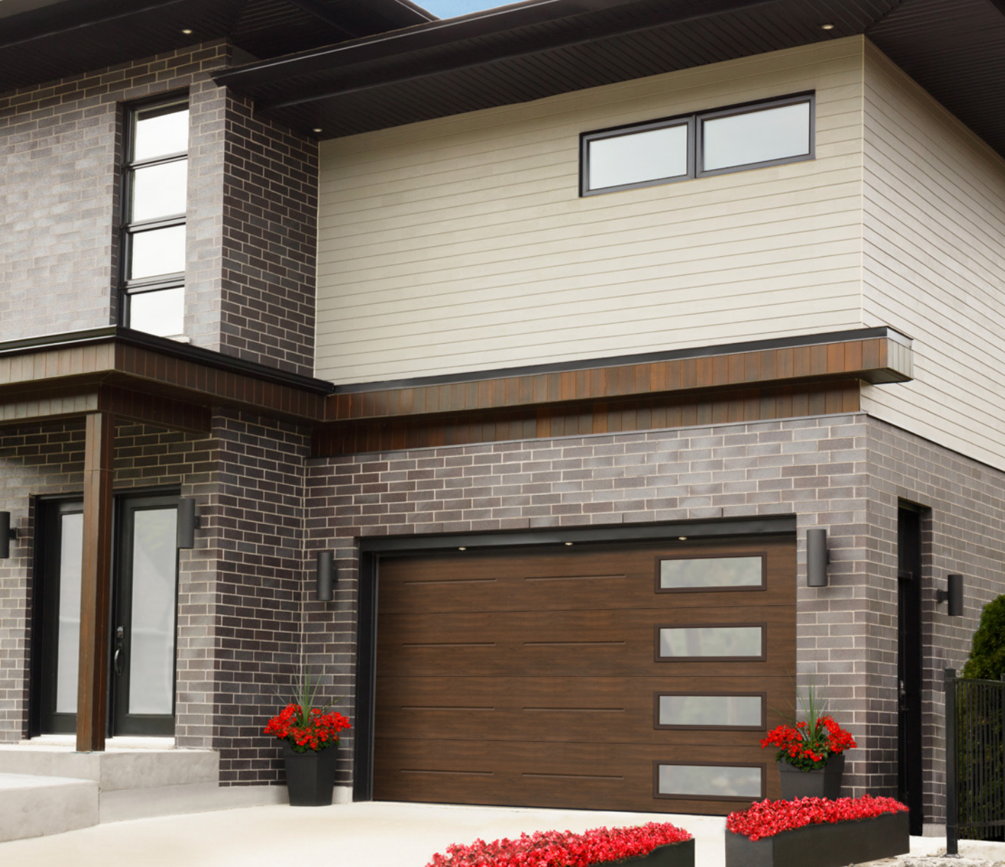 Extérieur d'une maison moderne à deux étages avec revêtement en briques et beige, porte de garage marron et fleurs rouges à l'avant.