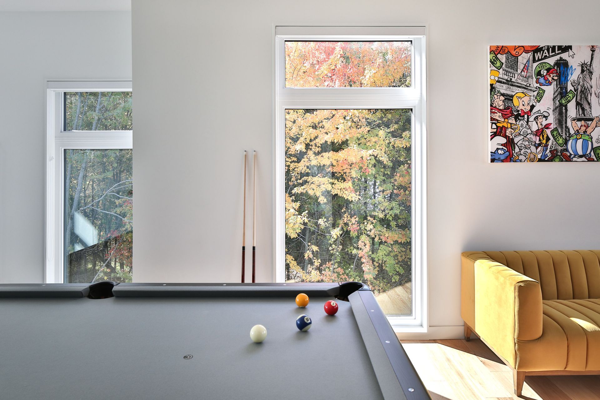 Une table de billard dans un salon lumineux avec des fenêtres, des tableaux et un canapé beige.