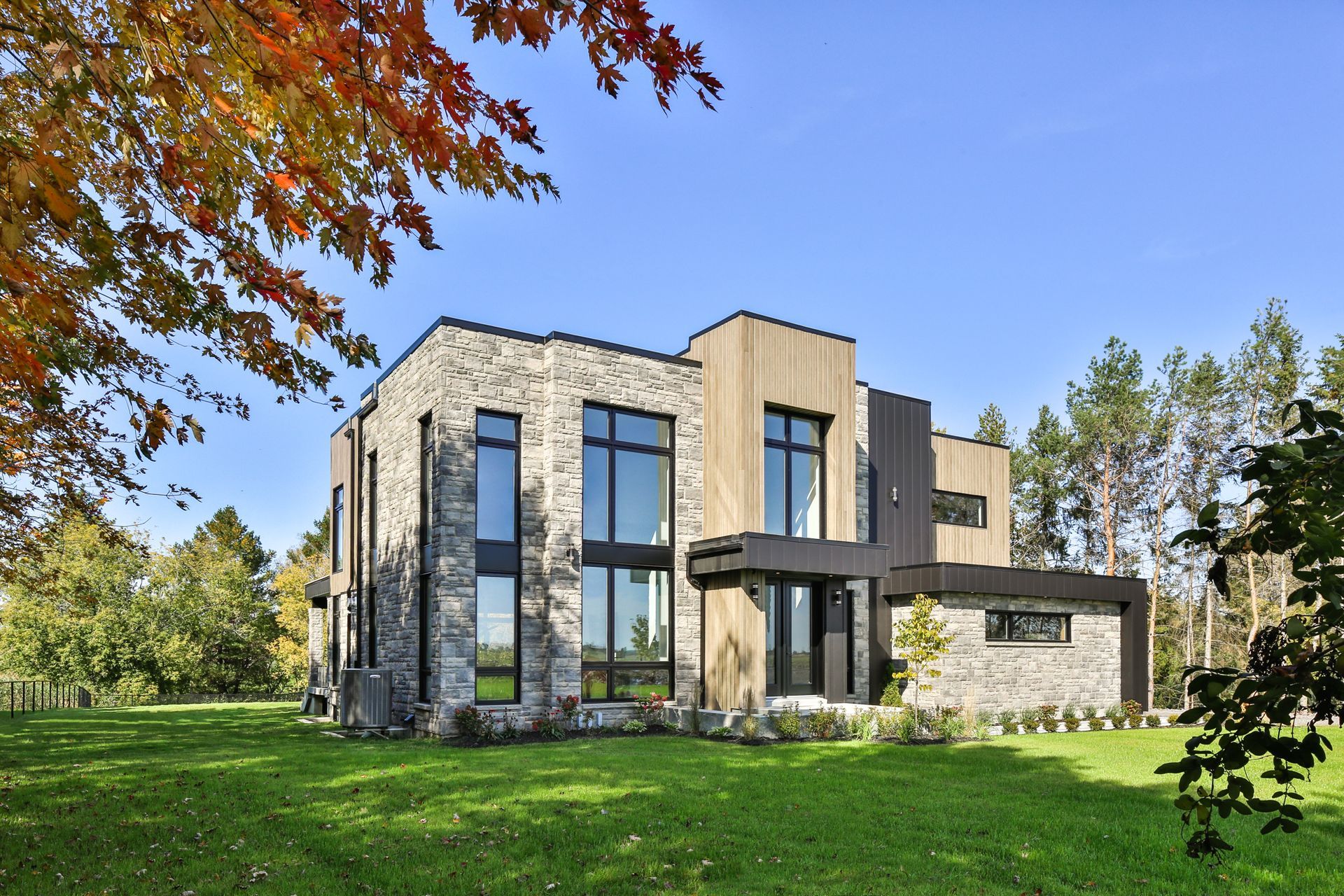Modern stone-and-glass house with large windows, a curved balcony, and a green lawn under a blue sky