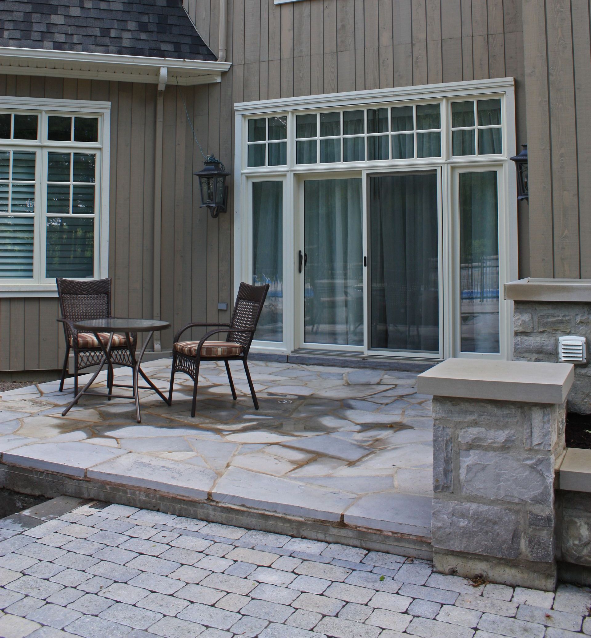 Terrasse avec deux chaises et une table en métal à l'extérieur d'une maison beige avec des portes coulissantes en verre et un dallage en pierre.