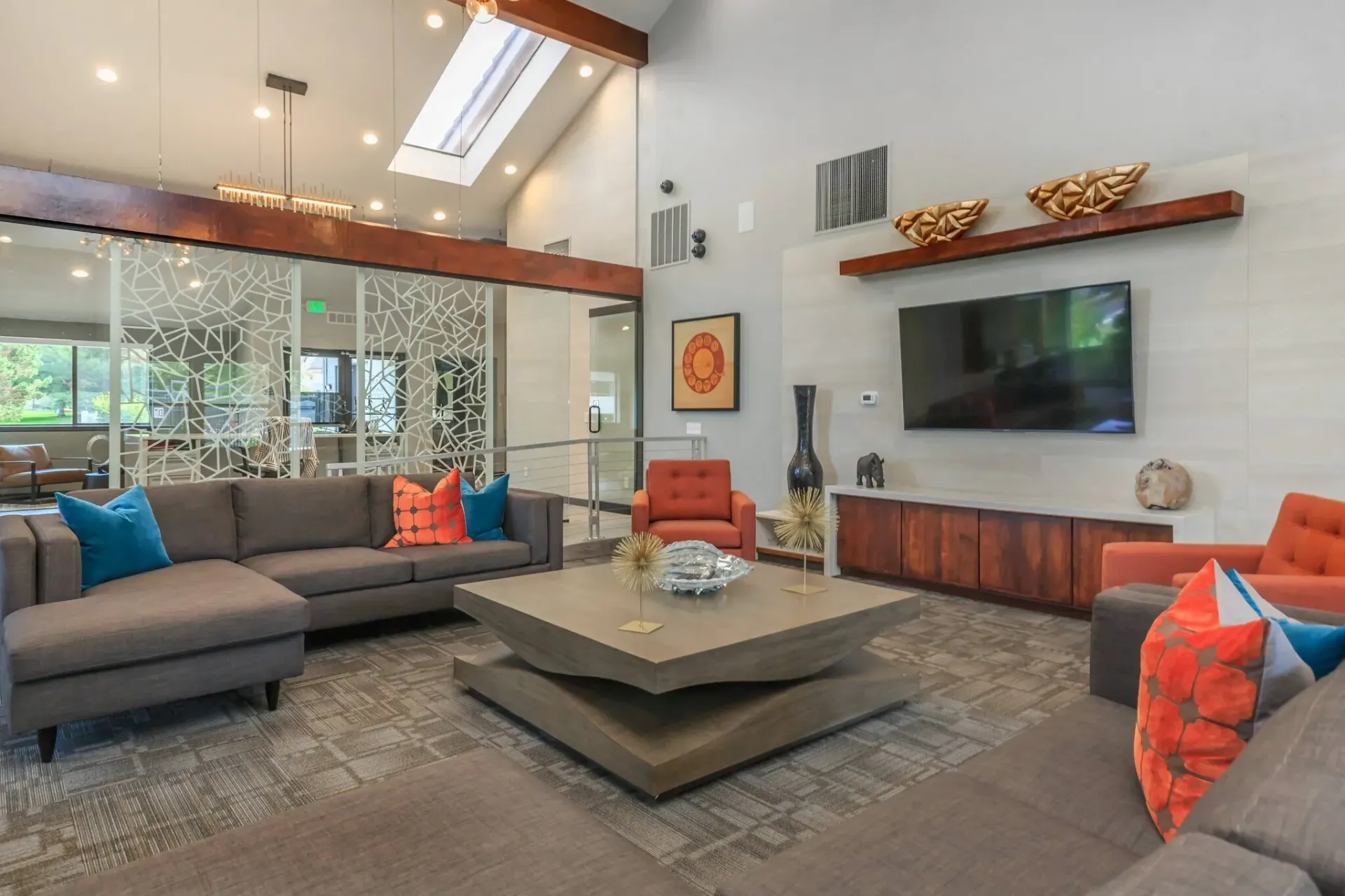 Modern living room with gray sofas, orange pillows, a wall TV, and a central coffee table beneath pendant lights.