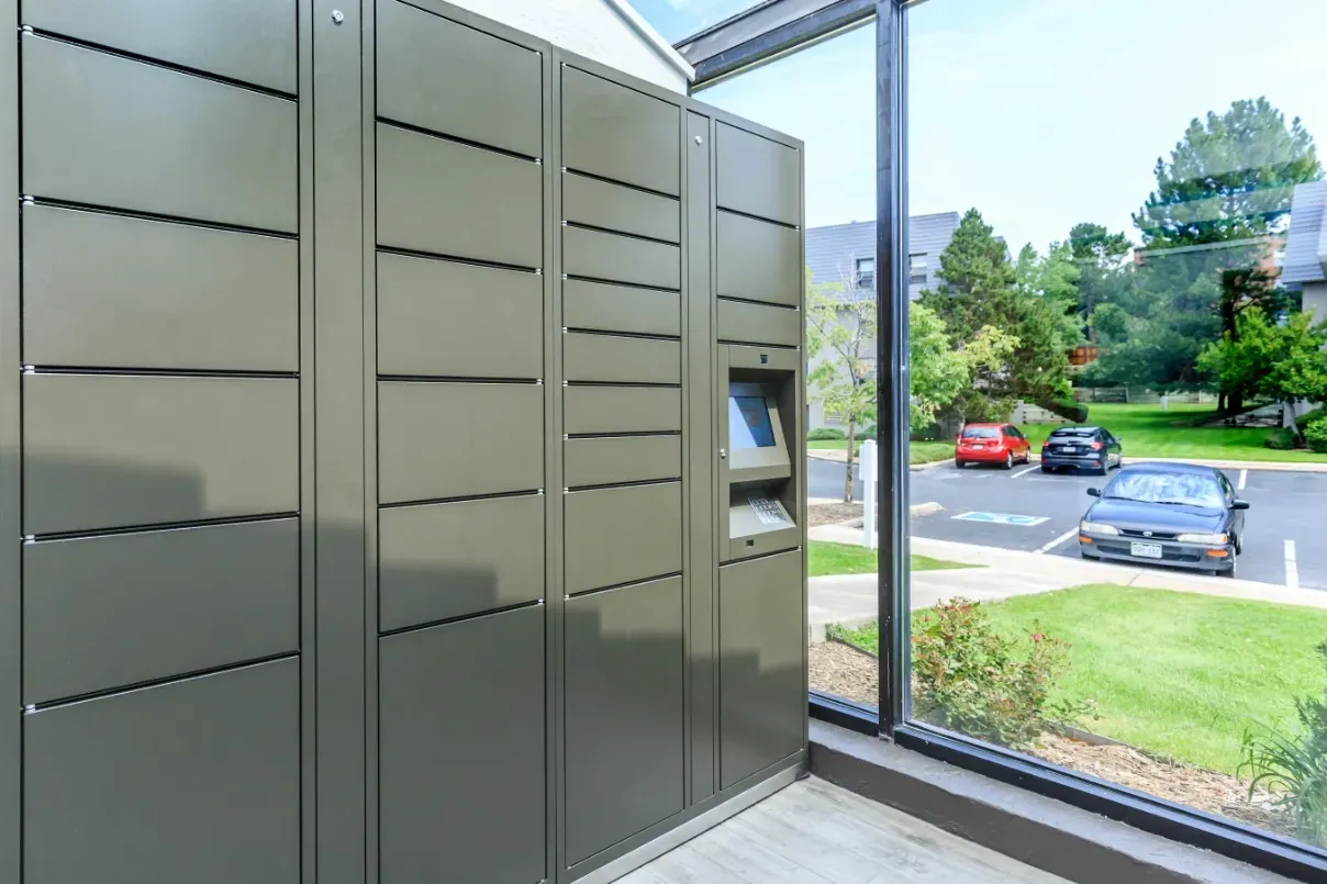 Secure package lockers with digital interface, viewed from inside a building through a large window.